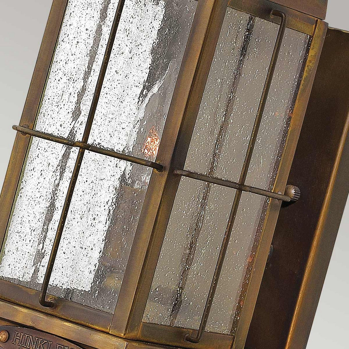 A close-up of the Bingham 1 Light Outdoor Small Wall Lantern in brown showcases its bronze finish with nautical influences, clear textured glass panels, and a visible bulb. This vintage design features a rectangular frame supported by vertical metal bars.