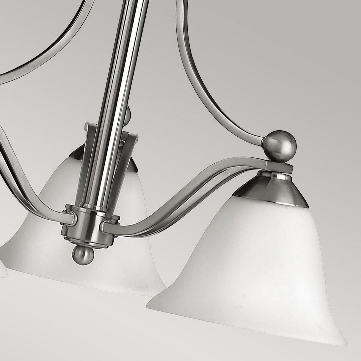 A close-up of the Bolla 3 Light Chandelier in brushed nickel showcases its smooth, curved metal arms and frosted white glass shades, set against a neutral gray background. This lighting fixture beautifully highlights a blend of contemporary and classic elements.