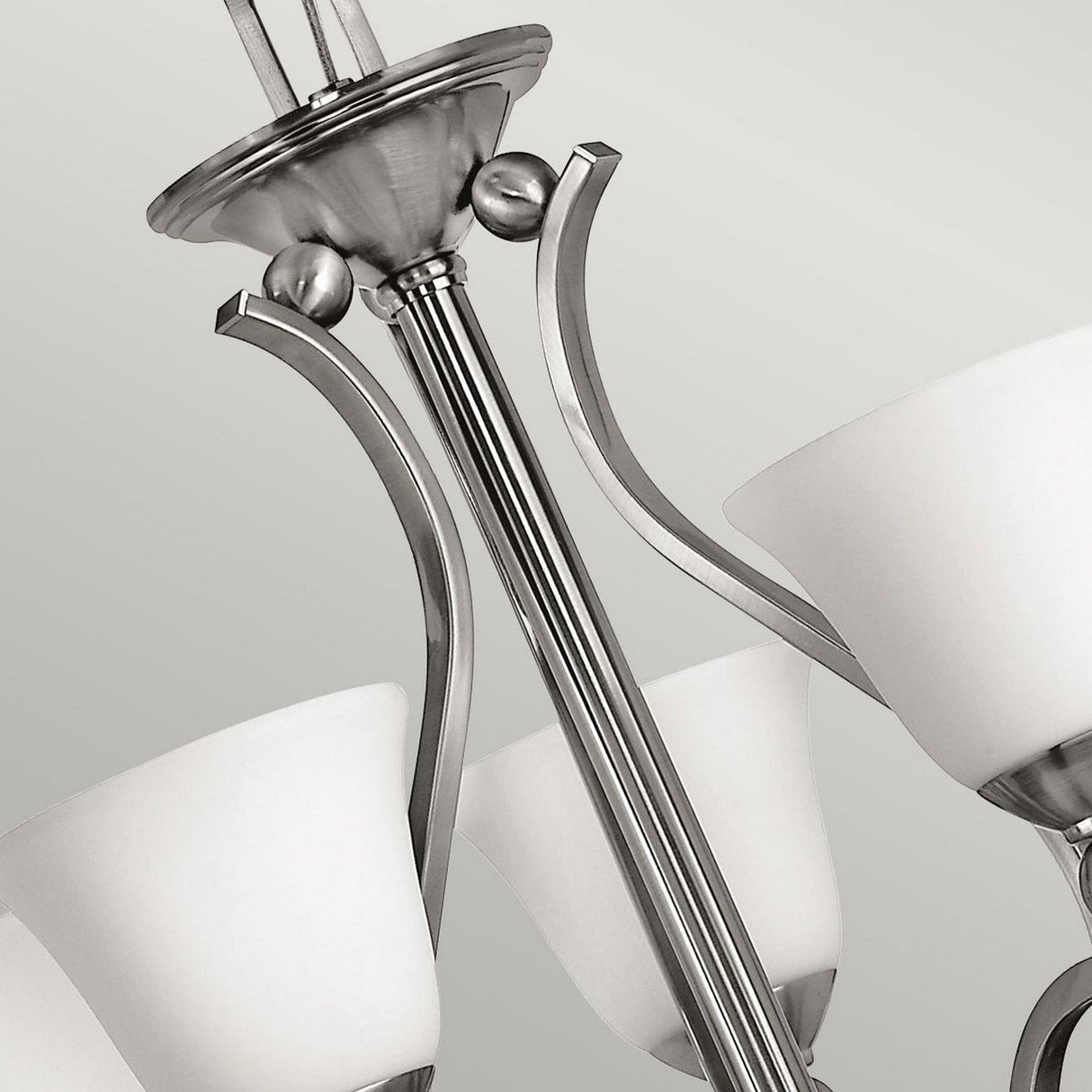 Close-up of the stylish Bolla 5 Light Chandelier - Brushed Nickel, featuring several white glass shades. Its smooth, curved arms elegantly link the shades to a central metal stem. The backdrop is a simple, light gray color.