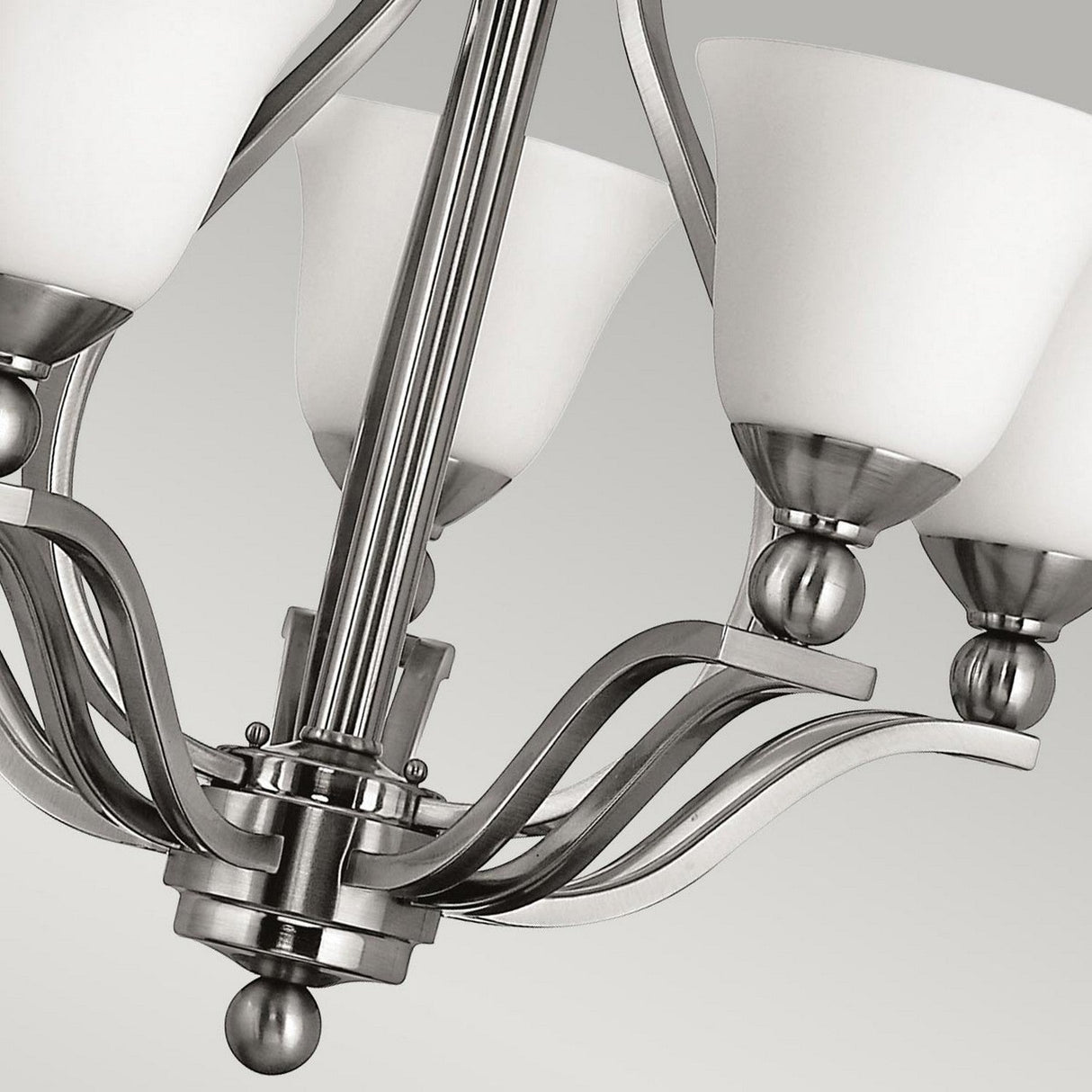 A close-up of the Bolla 5 Light Chandelier - Brushed Nickel highlights its five white glass shades and brushed nickel arms set against a neutral background. This elegant fixture showcases stylish curved metal accents, perfectly embodying modern sophistication.