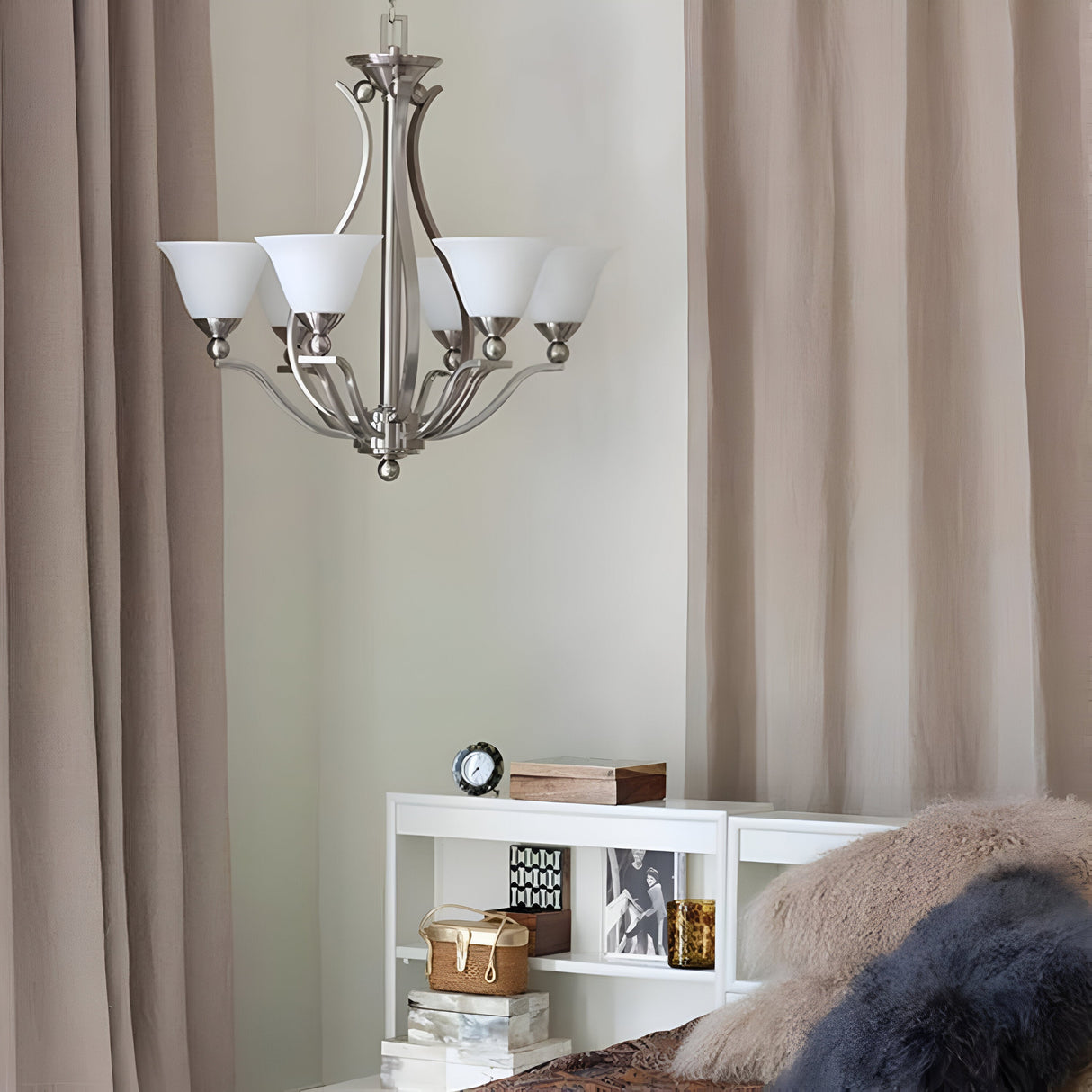 A modern bedroom interior showcasing an elegant Bolla 5 Light Chandelier in Brushed Nickel and beige curtains. The white nightstand, adorned with books, a small clock, and decorative items, complements the plush pillows on the bed for a cozy touch.