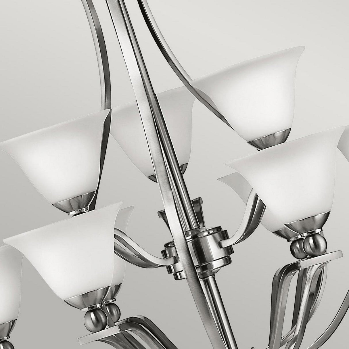 The Bolla 9 Light Chandelier - Brushed Nickel showcases a modern design with a sleek frame and six elegant, bell-shaped glass shades. Its symmetrical arrangement enhances any space with a touch of sophistication against a soft gray background.