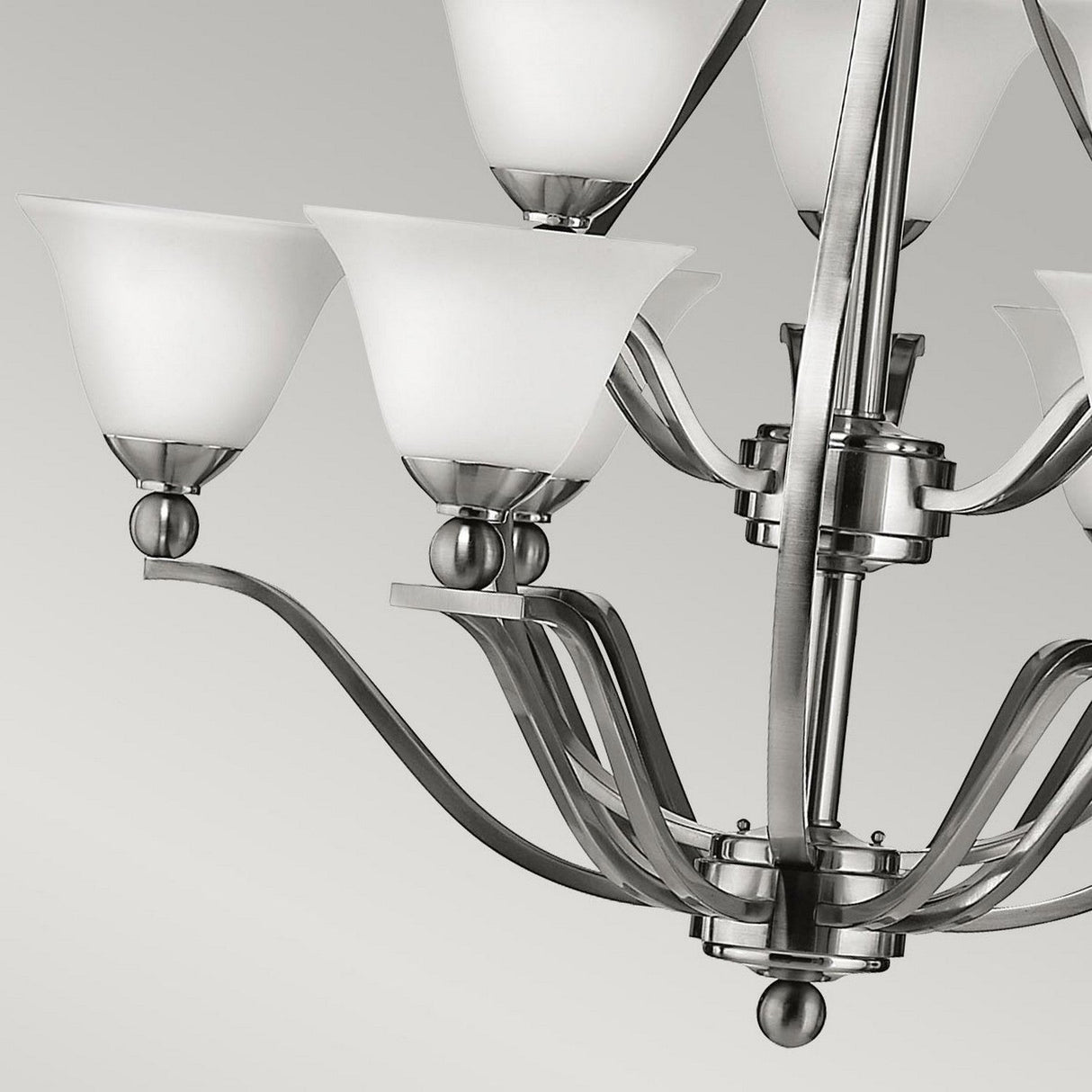 The Bolla 9 Light Chandelier - Brushed Nickel is displayed against a neutral gray background, highlighting eight bell-shaped white glass shades and a silver metal frame. The elegant, curved arms and central stem are accentuated by its sleek brushed nickel finish.