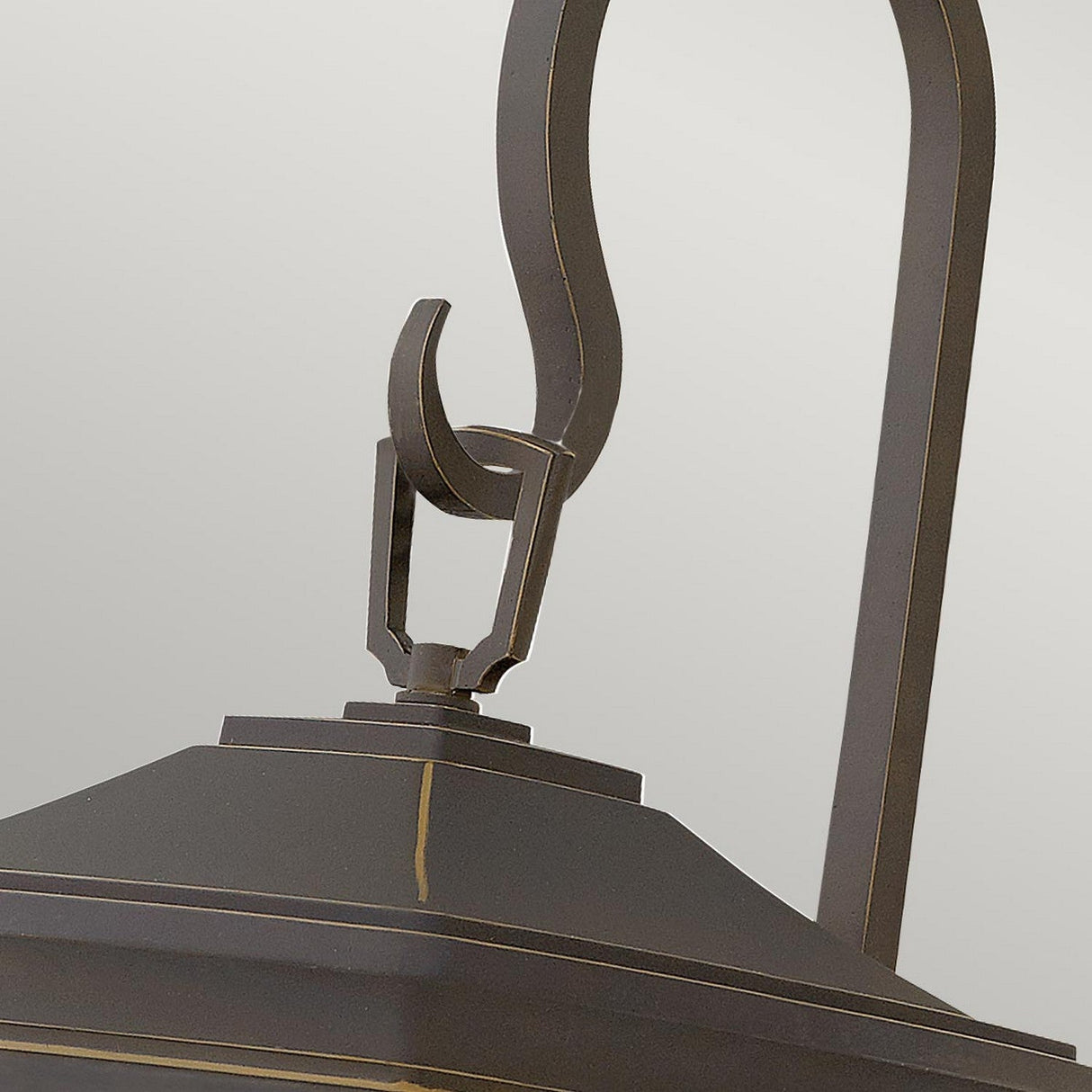 The Bromley 3 Light Outdoor Large Wall Lantern in bronze features a durable aluminum construction with an elegant geometric design topped by a curved hook. Subtle gold accents enhance its sophisticated appearance against a plain gray backdrop.