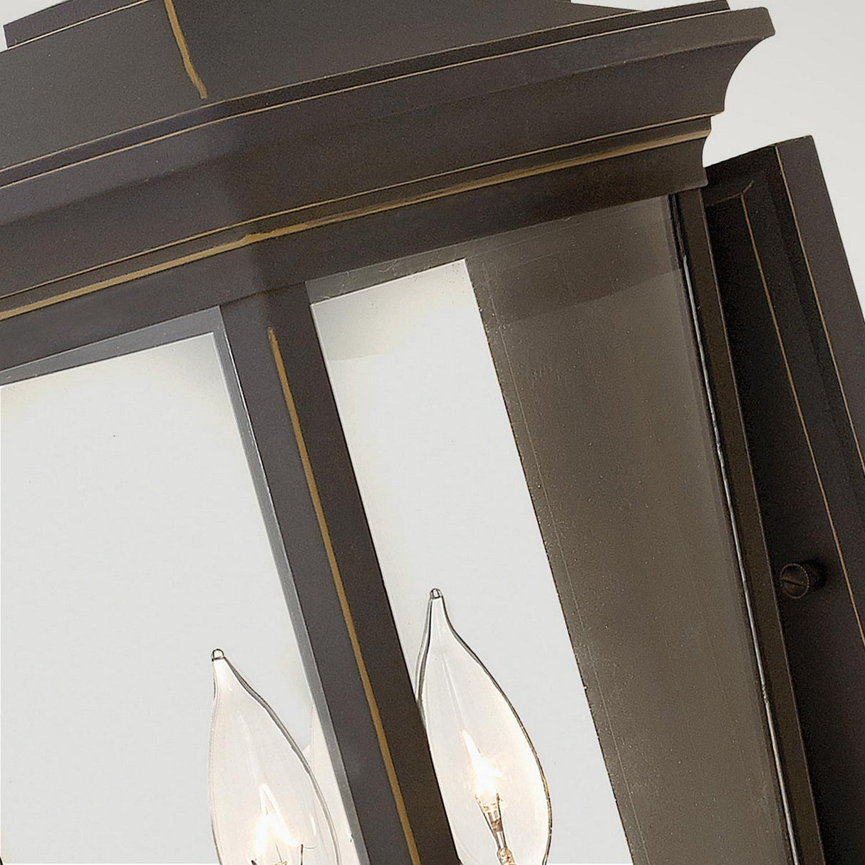Close-up of the Bromley 3 Light Outdoor Large Wall Lantern featuring a vintage style with a black metal frame and clear glass panes. Inside, two candle-shaped light bulbs are visible. This lantern is crafted from durable aluminum, comes in a bronze finish, and is mounted against a light-colored background.