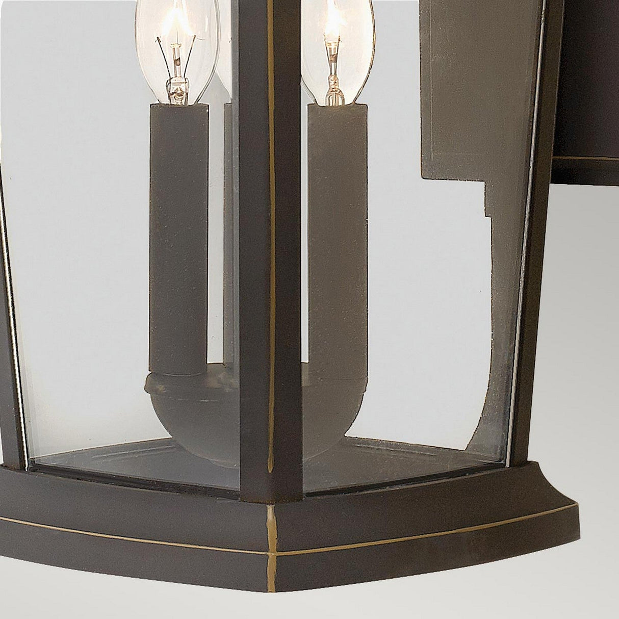 A close-up view of the Bromley 3 Light Outdoor Large Wall Lantern in bronze showcases its black metal and glass construction. The lantern houses two clear, elongated bulbs within its durable aluminum structure, featuring a geometric design with straight edges, all set against a plain gray background.