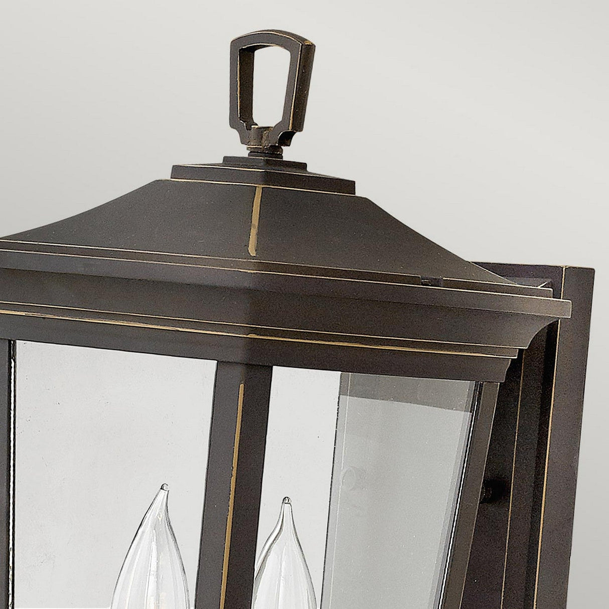 Close-up of the Bromley 2 Light Outdoor Medium Wall Lantern - Bronze. This lantern showcases glass panels and a classic black metal appearance. It features a small geometric handle on top, while its traditional design is highlighted by visible candle-shaped lightbulbs inside, perfectly complemented by an elegant bronze finish.