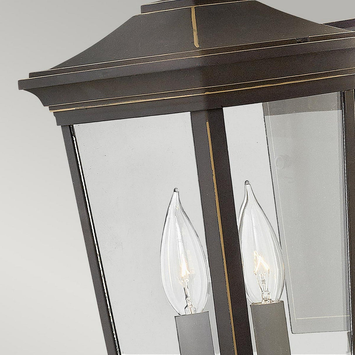 This close-up showcases the Bromley 2 Light Outdoor Medium Wall Lantern in bronze, featuring two candle-shaped light bulbs. It boasts a traditional design with a dark finish, highlighted by delicate golden and bronze accents, all set against a soft gray background.