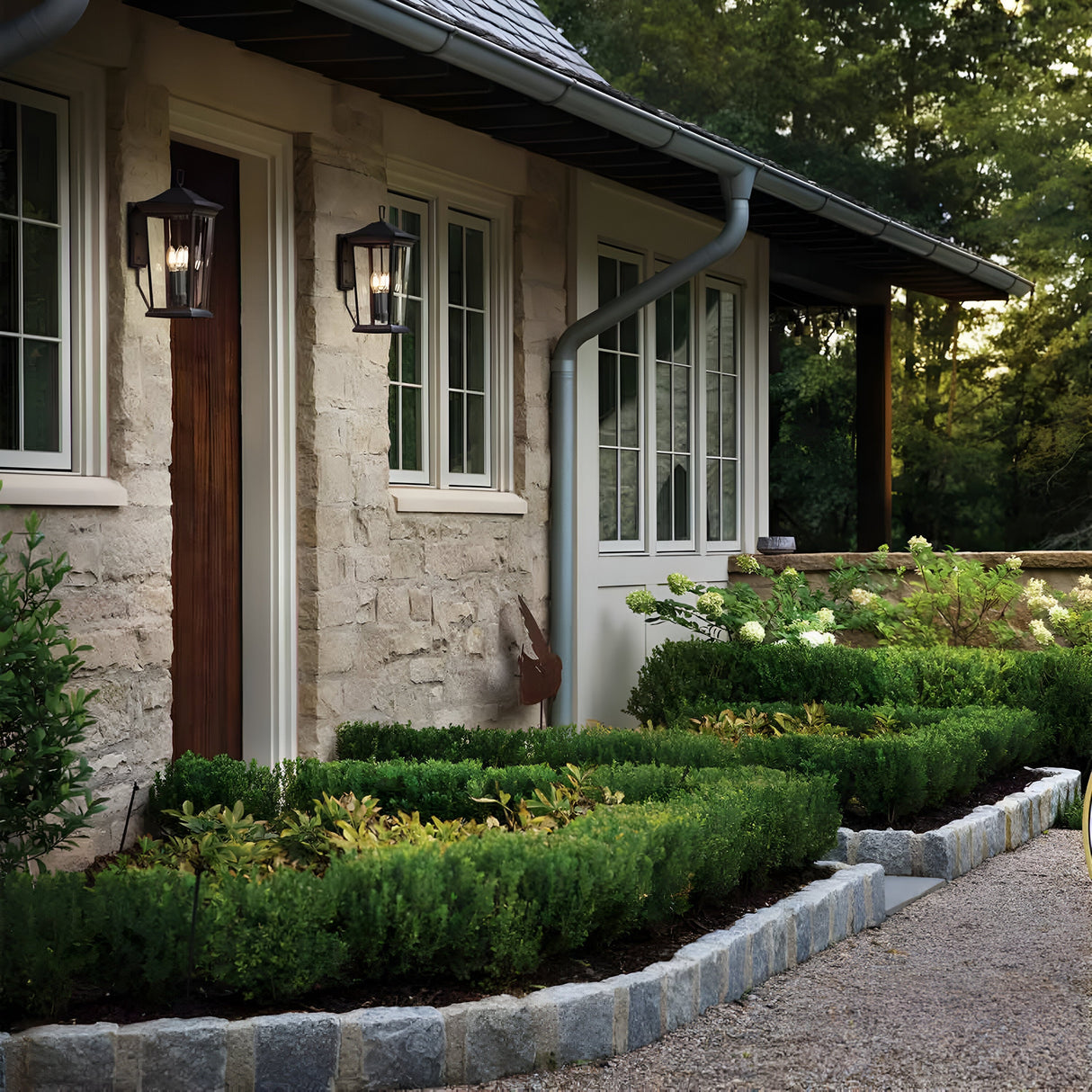 A charming stone cottage with a traditional design features two Bromley 2 Light Outdoor Medium Wall Lanterns in a bronze finish flanking the wooden door. Trimmed hedges and shrubs line the gravel path to the entrance. The facade boasts multiple windows, while lush trees stand majestically in the background.