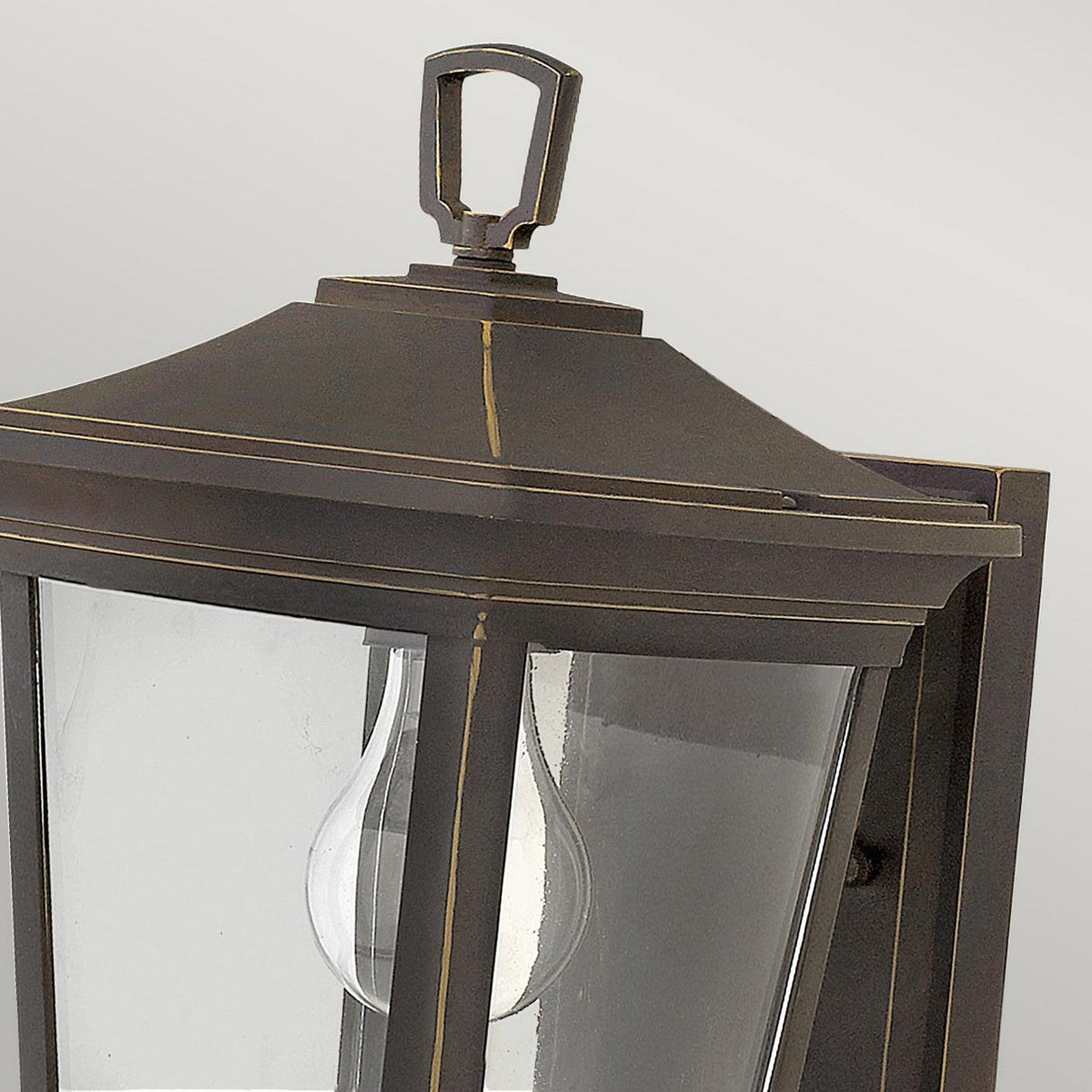 Close-up of the Bromley 1 Light Outdoor Small Wall Lantern - Bronze, crafted from die-cast aluminum with clear glass panels showcasing a visible light bulb inside. The lantern features a traditional design with an ornamental top detail and boasts an elegant bronze finish.