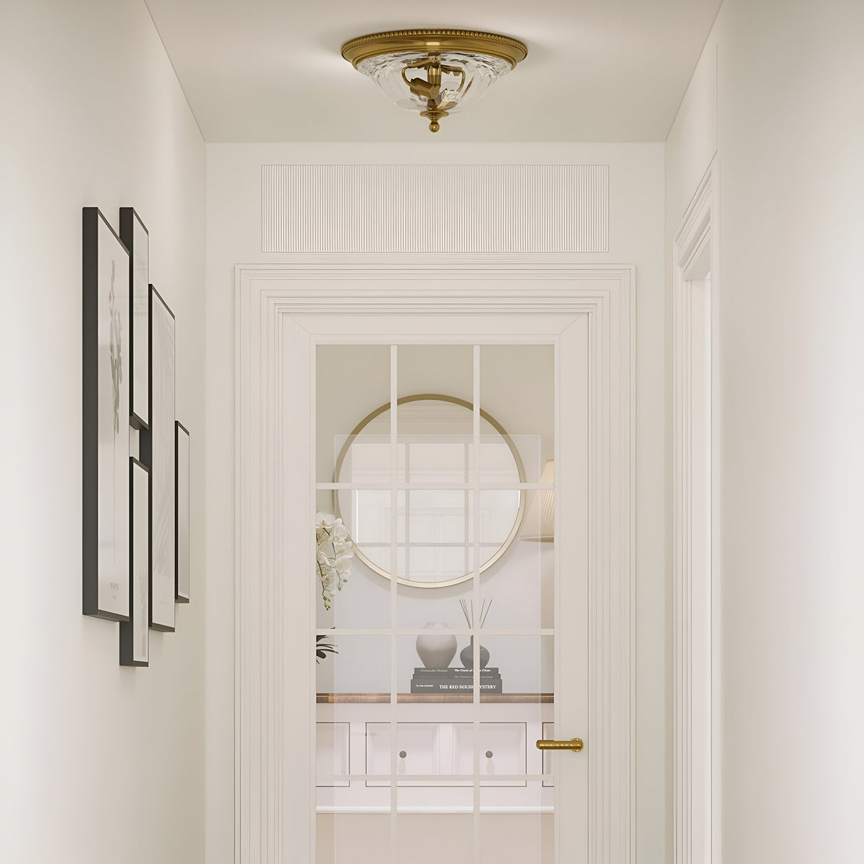 A well-lit hallway with white walls and framed artworks on the left exudes New England decor charm. The door, featuring glass panes, offers a view of a round mirror and books on a console table. Positioned above and enhancing the elegance of the setting is the Cambridge 2 Light Flush in Burnished Brass.