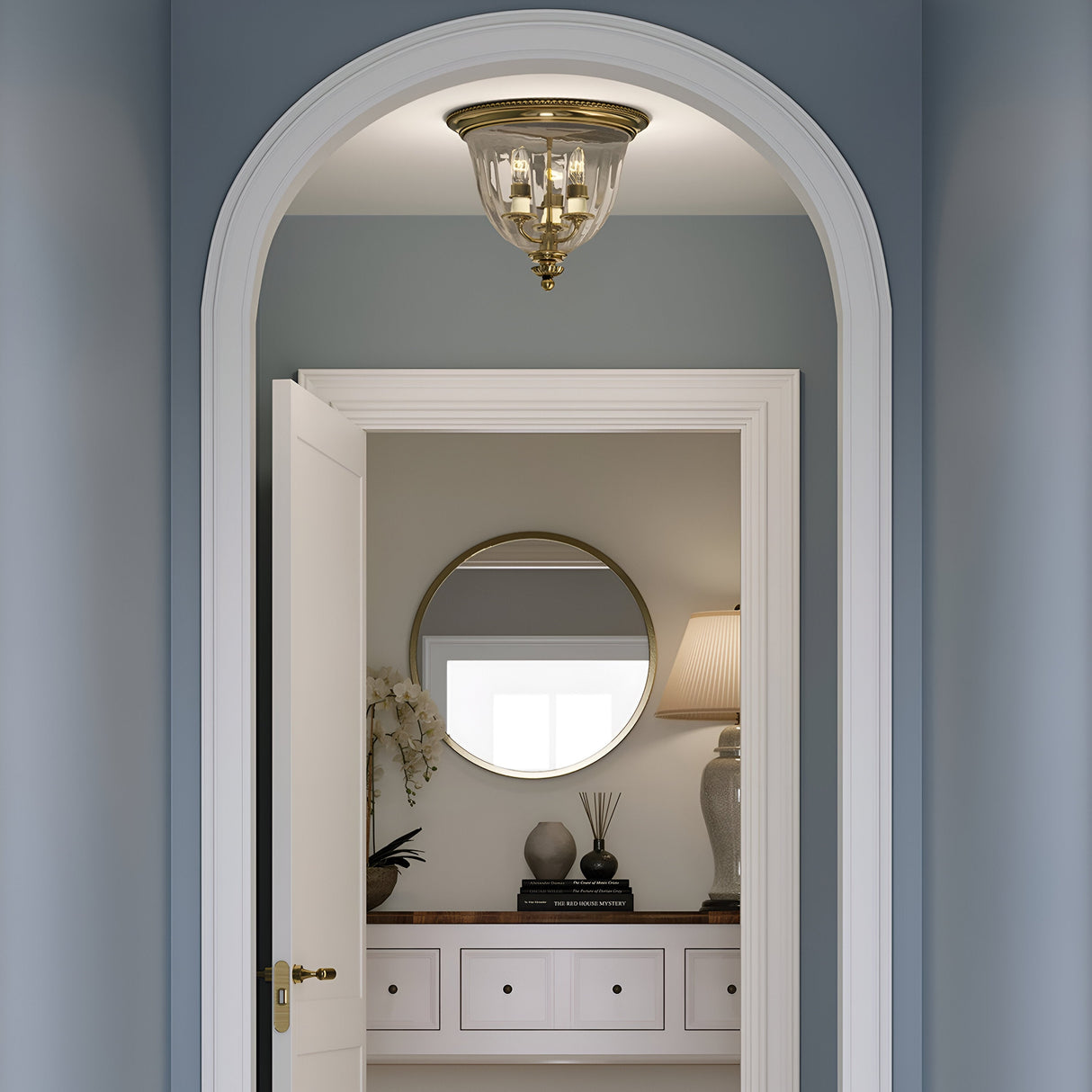 A hallway painted in light blue leads to a well-lit room. The arched doorway features a slightly ajar white door. Inside, there's a round mirror on the wall, accompanied by a lamp and decor on a console table, adding a touch of New England style. A Cambridge 3 Light Flush - Burnished Brass fixture gleams above the arch.
