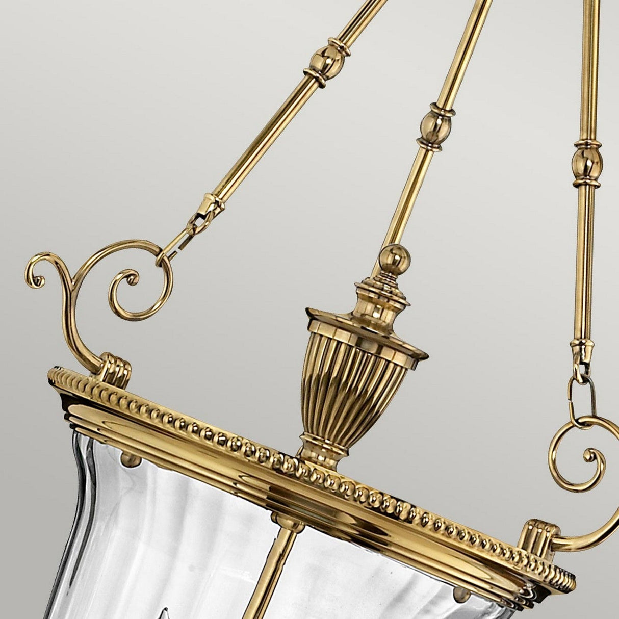 Close-up of a Cambridge 3 Light Chandelier - Burnished Brass. This elegant fixture, crafted from solid brass, features ornamental detailing and intricate metalwork with curved accents and a decorative finial. The glass enclosure stands out against the plain, neutral background.
