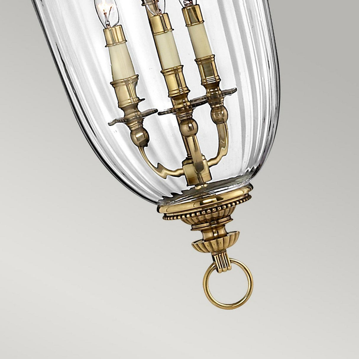This sophisticated chandelier, the Cambridge 3 Light Chandelier - Burnished Brass, showcases a burnished brass finish and solid brass craftsmanship. It includes three candle-style bulbs encased in ribbed glass with intricate detailing and a circular hanging ring at the bottom.