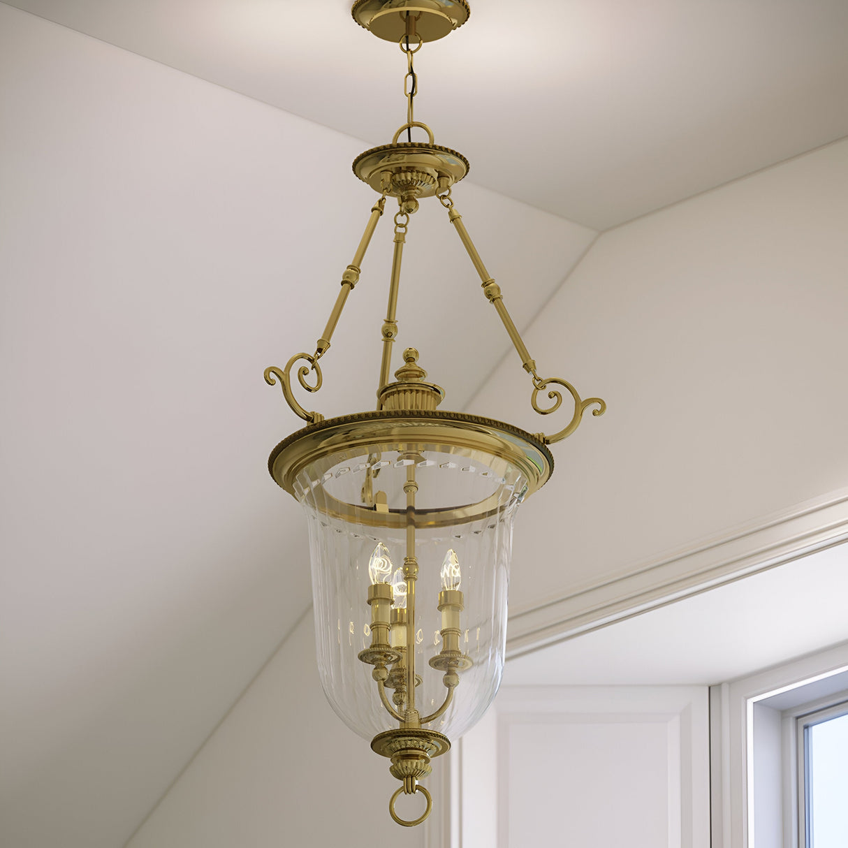 The Cambridge 3 Light Chandelier - Burnished Brass gracefully hangs from the ceiling, showcasing its elegant design and solid brass construction. It holds three candle-style bulbs within a generous, clear glass shade, beautifully lighting up a room with white walls and an angled ceiling.