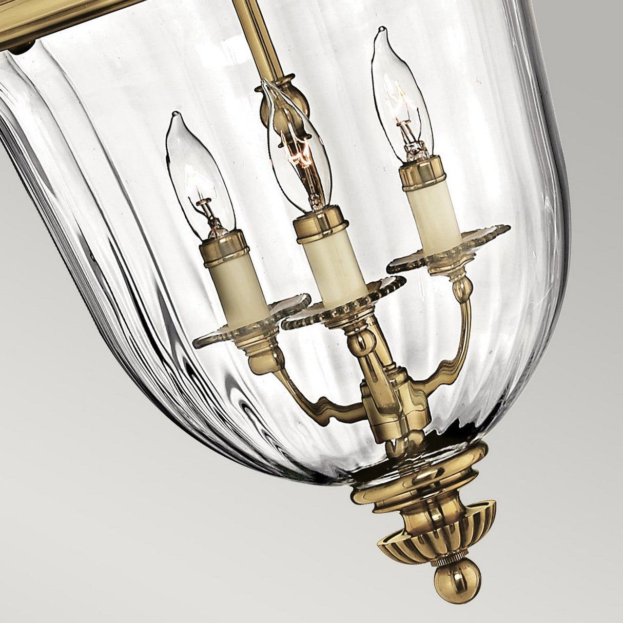 The Cambridge 3 Light Small Chandelier in Burnished Brass features an elegant design with three candle-shaped light bulbs encased in clear glass. It showcases intricate detailing and a classic New England style, beautifully contrasted against its light gray background.
