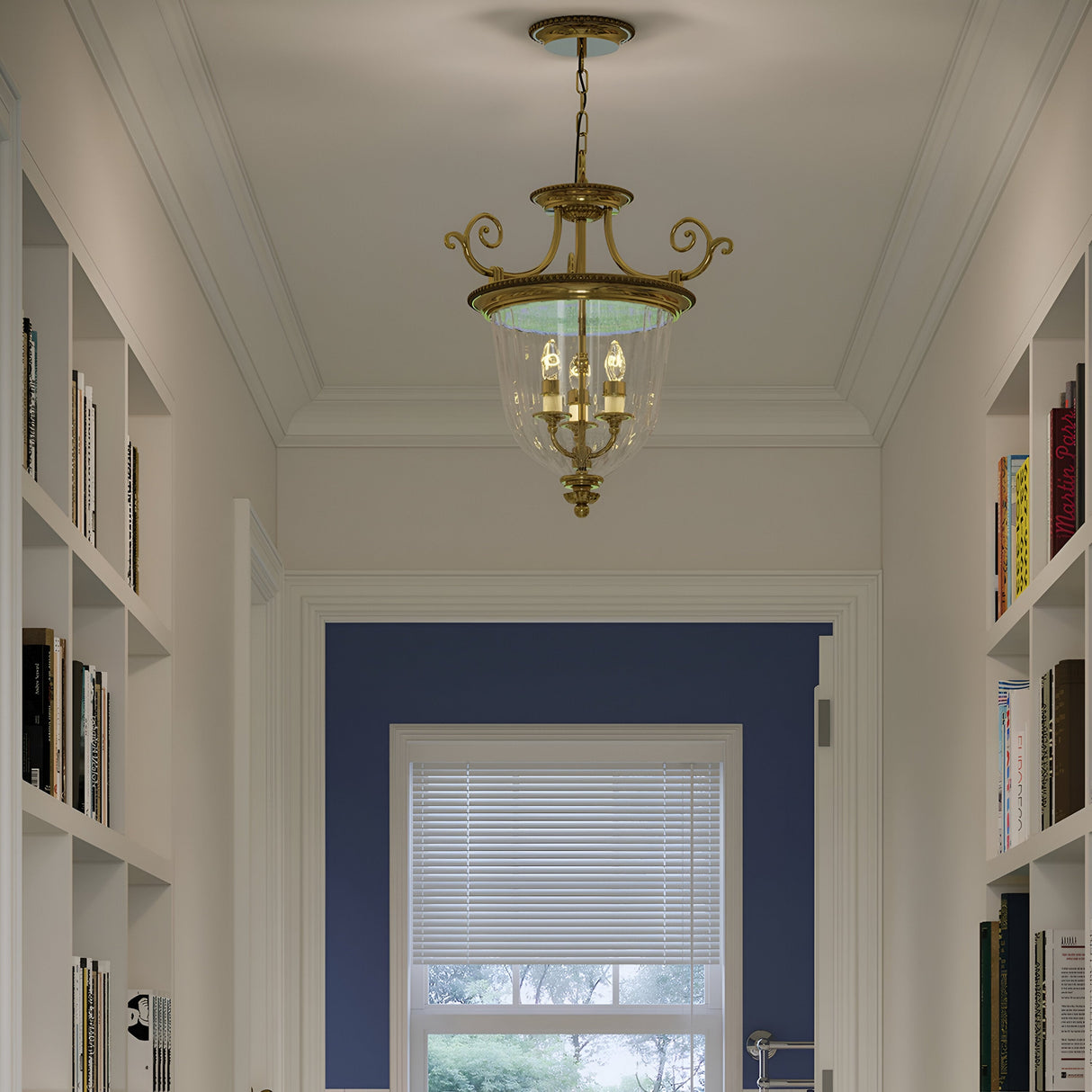 The hallway, echoing New England design, boasts white shelves brimming with books on either side. A large window at the end offers an outdoor view, partially covered by a white blind. Gracefully suspended from the ceiling is the Cambridge 3 Light Small Chandelier in Burnished Brass.