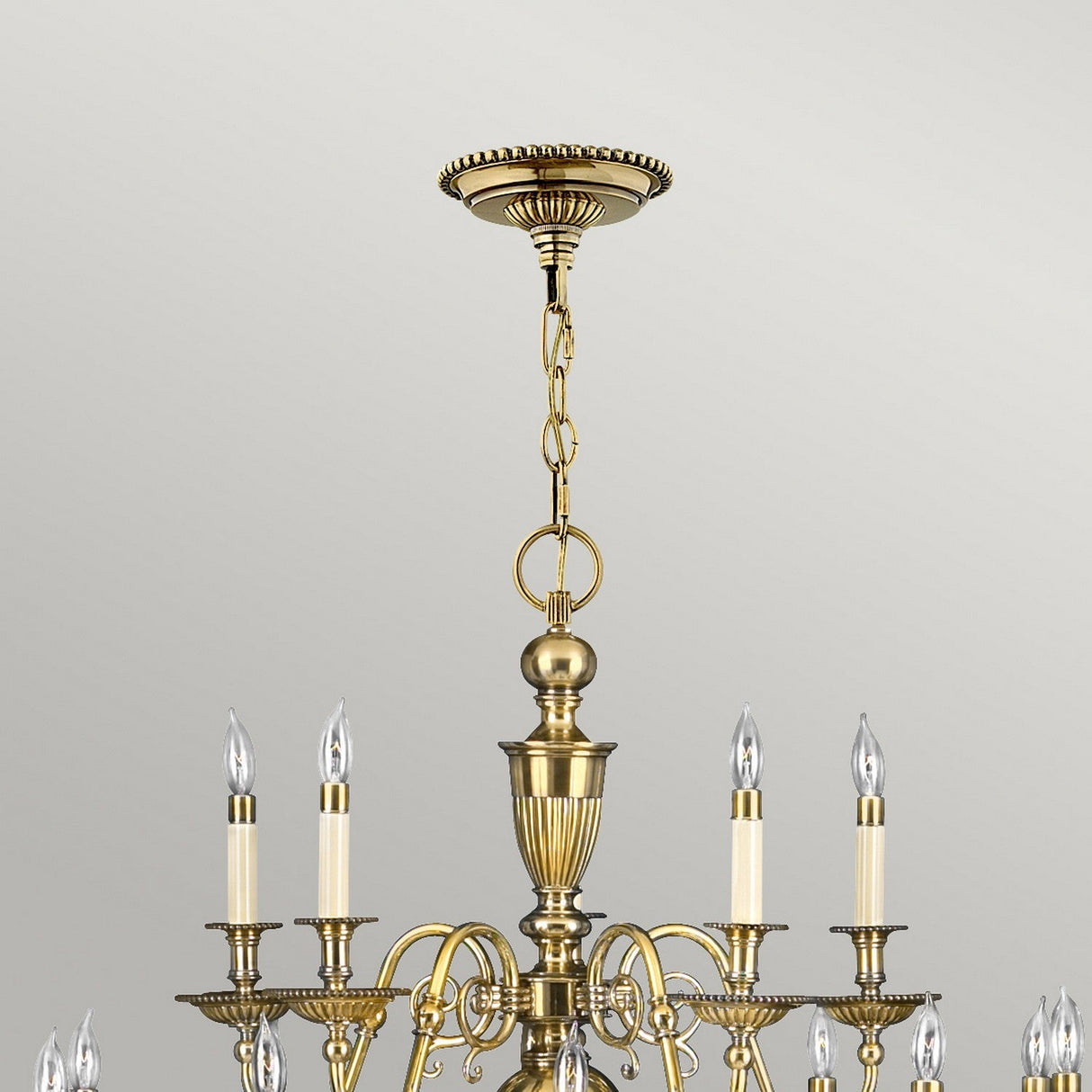 The Cambridge 25 Light Chandelier - Burnished Brass radiates New England elegance with its burnished brass finish. This ornate design includes multiple candle-shaped bulbs and decorative chain links, gracefully hanging from a detailed ceiling mount against a light gray background.