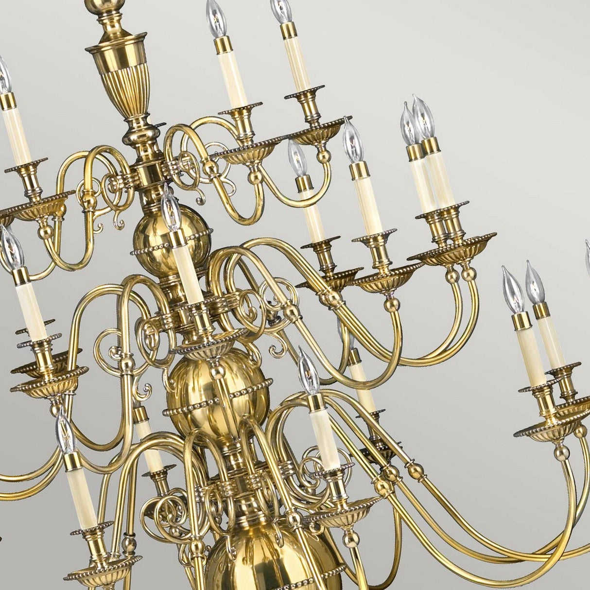 The Cambridge 25 Light Chandelier in burnished brass boasts an elegant New England design with its intricate scrolling and decorative details. Each arm holds a candle-shaped light bulb, creating a stunning contrast against a light gray backdrop.