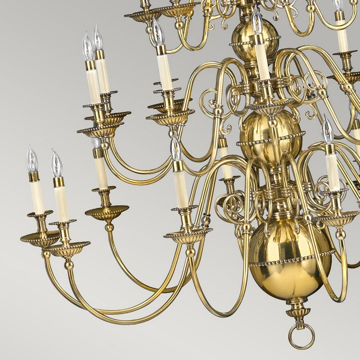 The Cambridge 25 Light Chandelier in Burnished Brass radiates New England elegance, featuring an intricate design. The swirling arms are adorned with candle-like bulbs, and its polished finish shines beautifully against a simple, light backdrop.