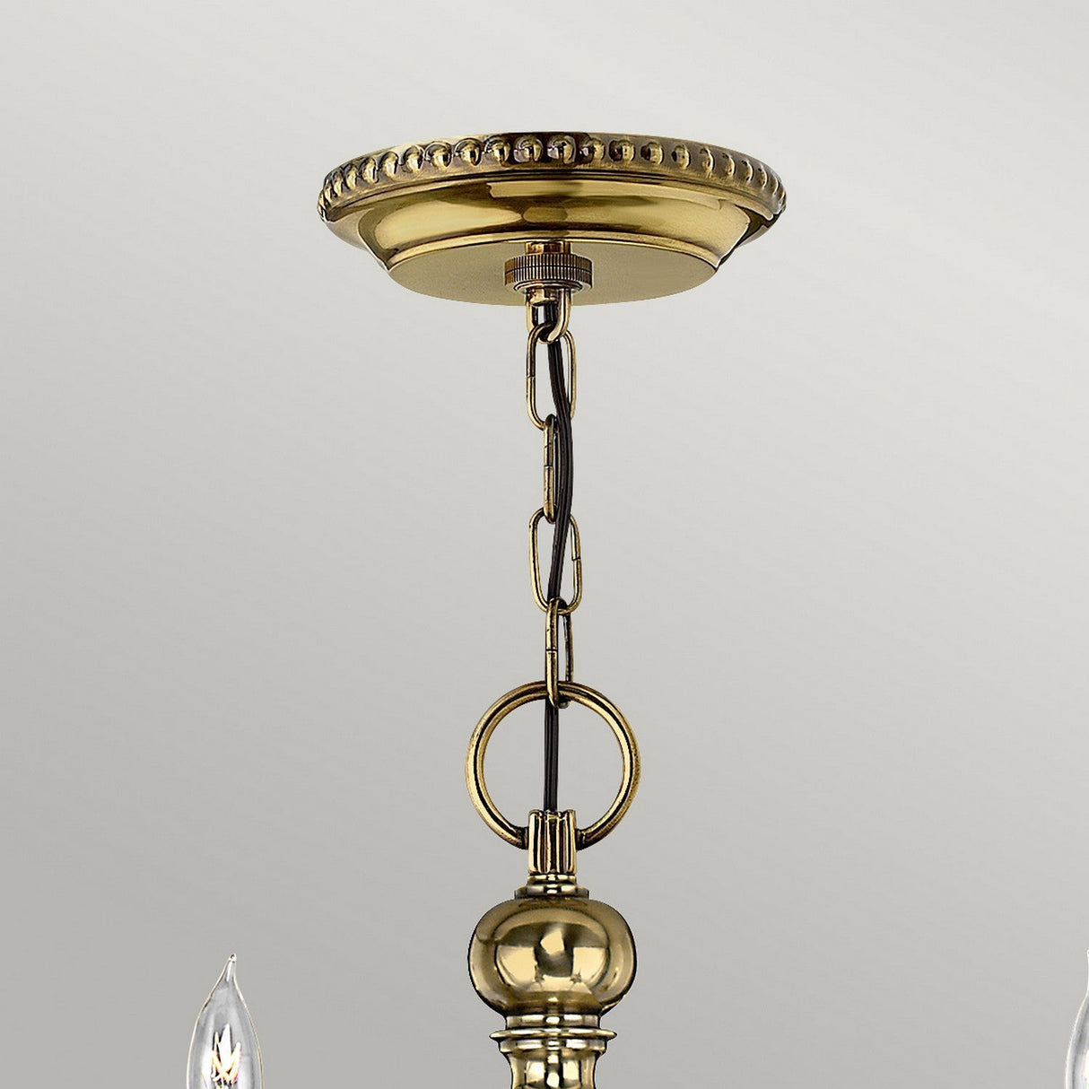 A close-up of the Cambridge 6 Light Chandelier in burnished brass showcases its ornate beaded ceiling plate. The fixture includes a decorative chain, a round central accent, and partially visible candle-like bulbs beneath. The elegant design is set against a plain gray background.