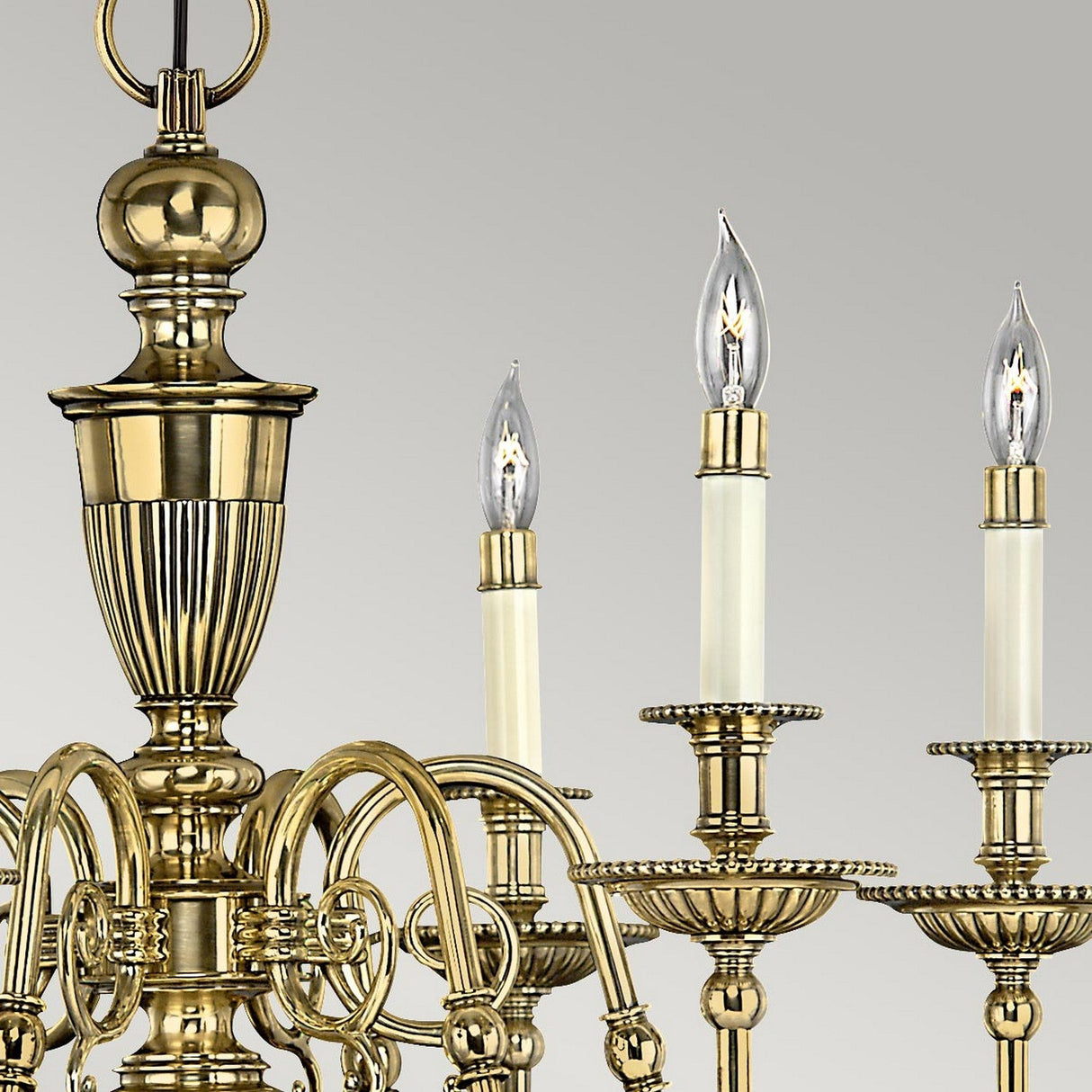 A close-up of the Cambridge 6 Light Chandelier - Burnished Brass reveals three candle-shaped bulbs and intricate detailing on its frame. The curved arms gracefully support optic glass shades, all set against a gray background.