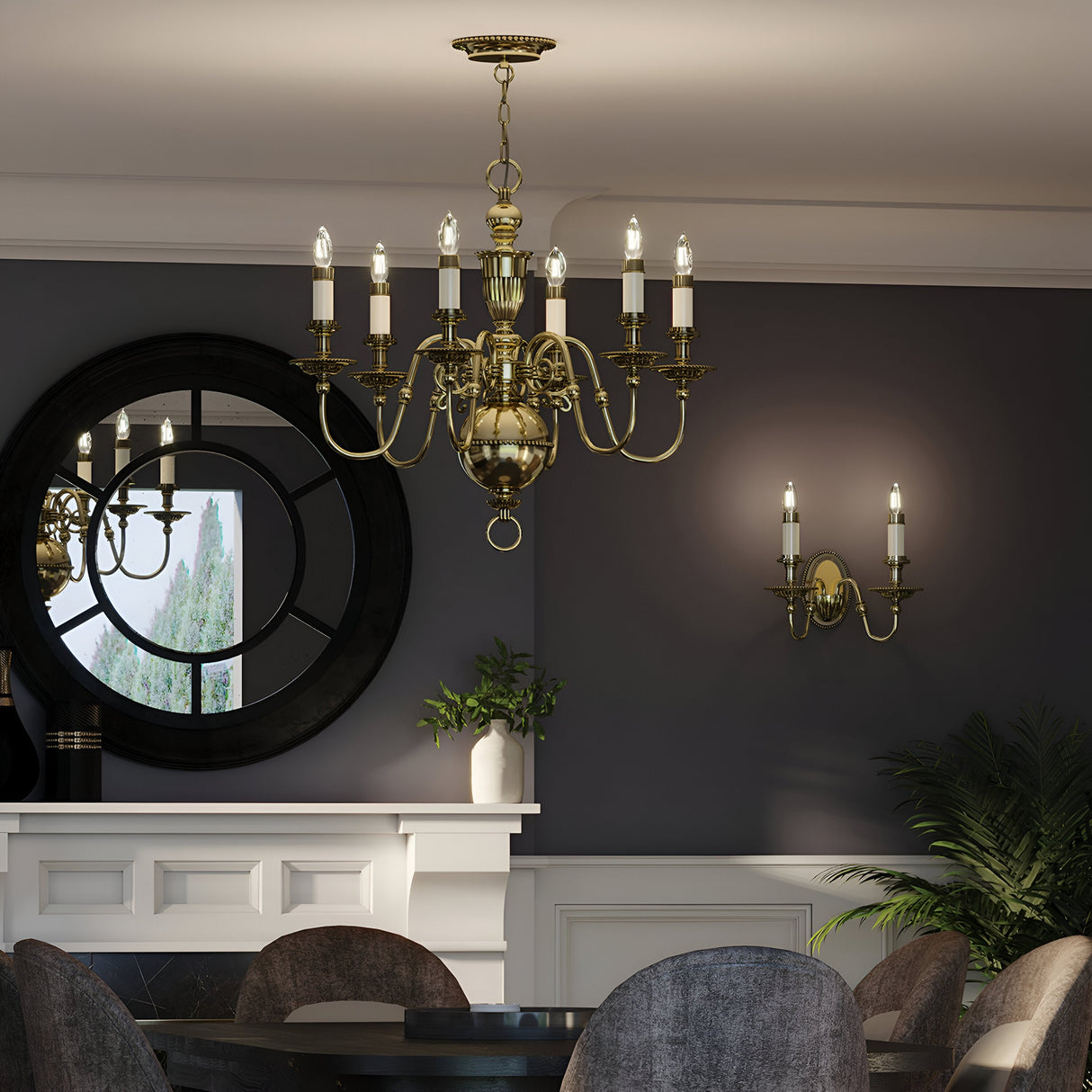 The sophisticated dining room is illuminated by a central Cambridge 6 Light Chandelier in burnished brass, featuring candle-like lights. A round table is surrounded by gray chairs, while a large mirror reflects the greenery. The dark walls are enhanced with matching candle-style wall sconces, also in burnished brass.