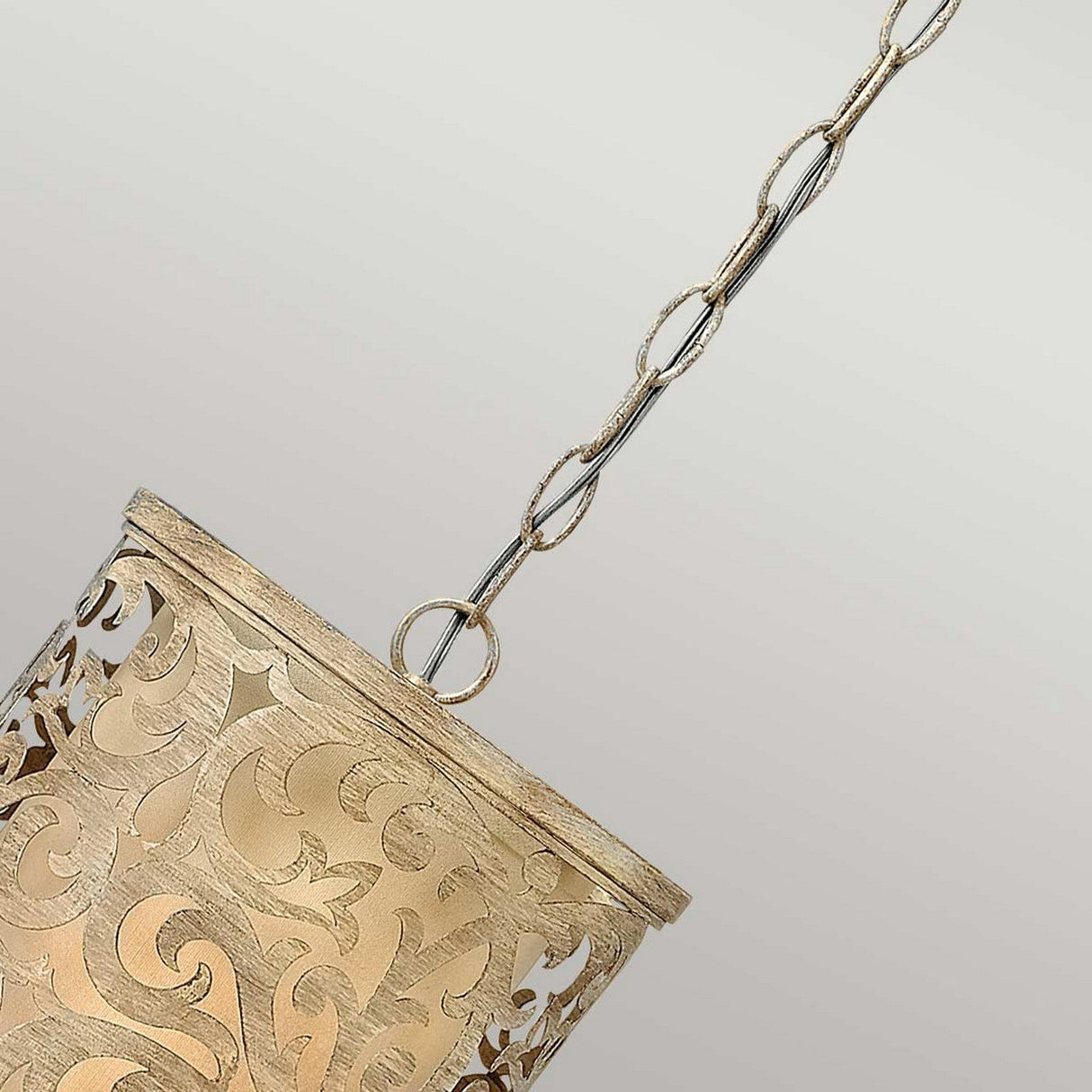The Carabel 1 Light Mini Pendant in Brushed Champagne showcases pierced metalwork with ornate swirling cutout patterns, featuring a gold-hued hanging lamp suspended by a decorative chain against a plain gray background.