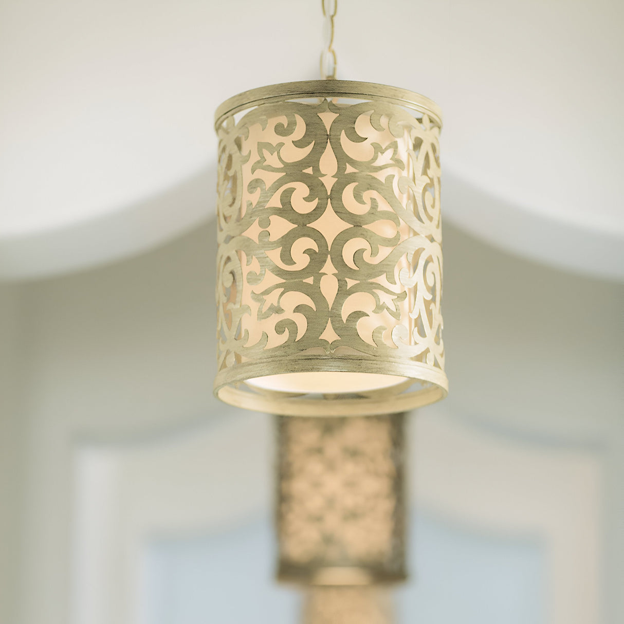 A Carabel 1 Light Mini Pendant in a brushed champagne finish hangs from the ceiling, showcasing its intricate pierced metalwork design and emitting a warm glow that highlights the ornate pattern. Another similar pendant can be seen slightly blurred in the background.