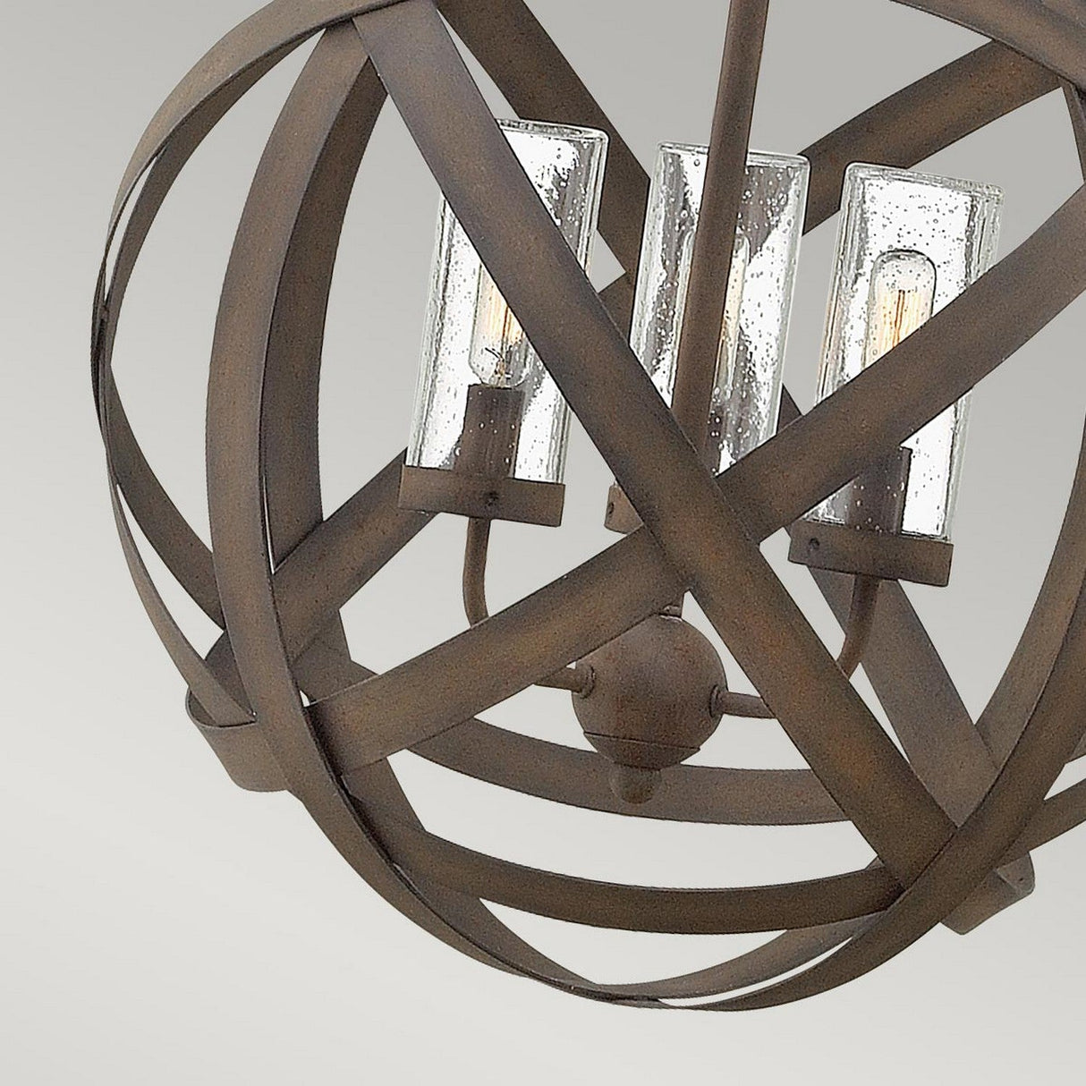 The Carson 3 Light Outdoor Chandelier - Vintage Iron features a unique orb-like design with interwoven metal bands in a vintage iron finish. The interior reveals three glass-enclosed light bulbs that contribute to its rustic charm and sophisticated industrial aesthetic, perfectly set against a soft, neutral gray background.