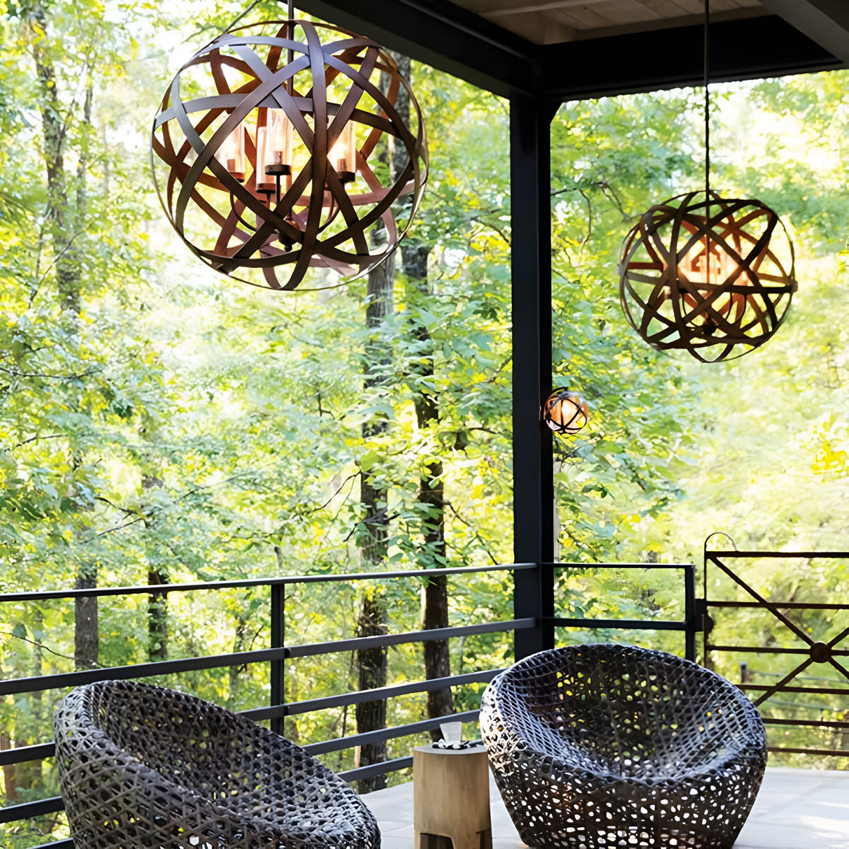 A wooden deck with round, woven chairs and a small side table exudes rustic charm. The stylish Carson 5 Light Outdoor Chandelier in Vintage Iron hangs overhead, resembling an elegant outdoor chandelier. The deck overlooks a lush, green forest, creating a serene outdoor setting.