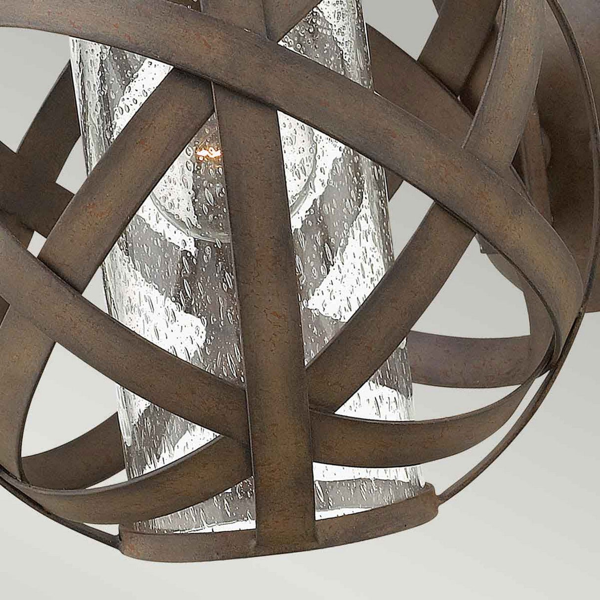 Close-up of the Carson 1 Light Outdoor Small Wall Lantern in Vintage Iron, featuring intersecting metal bands encasing a textured glass shade. A warm bulb inside emits a soft glow, complementing the rustic finish of the vintage iron.