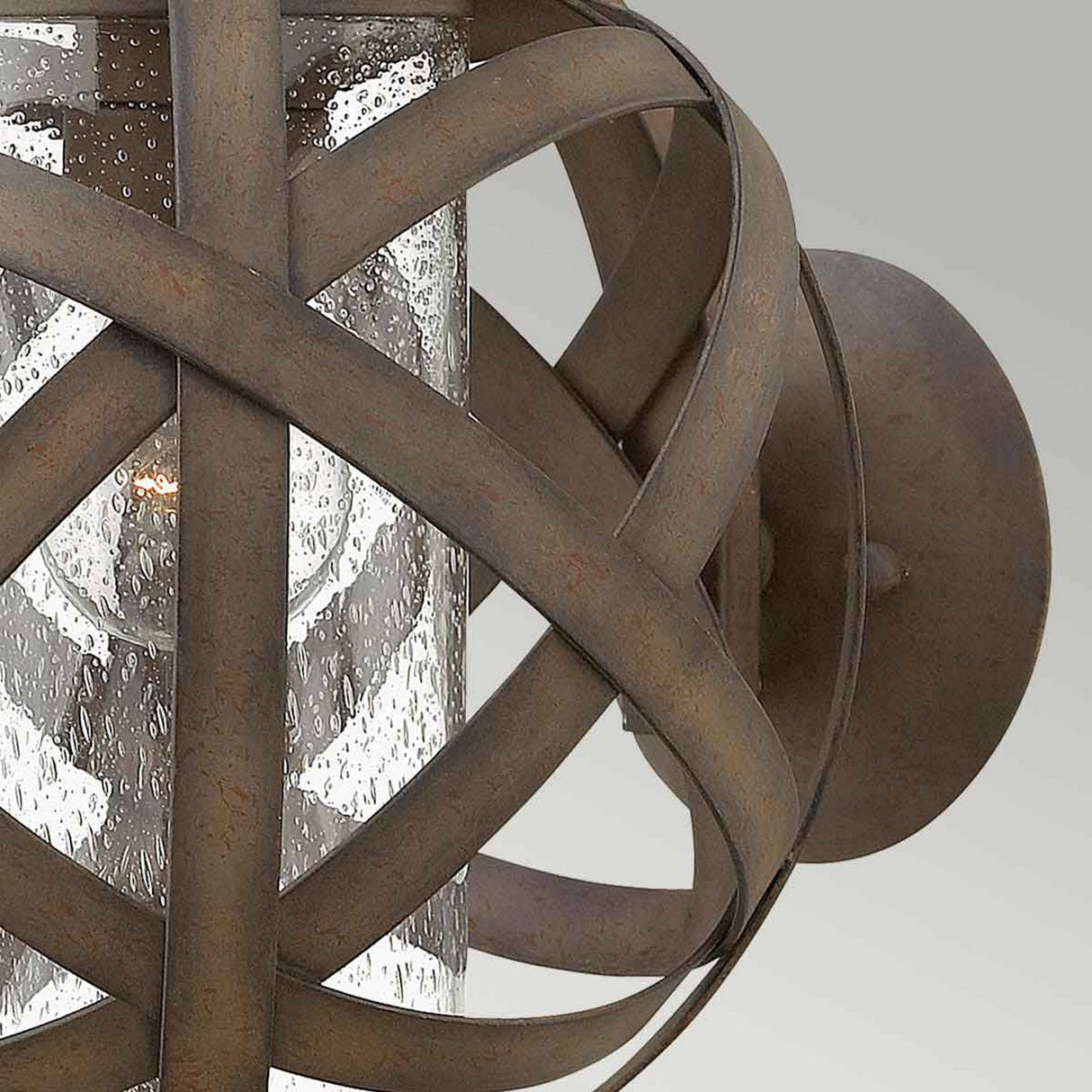 A close-up of the Carson 1 Light Outdoor Small Wall Lantern in Vintage Iron showcases its industrial design. This handcrafted lantern features a clear glass cylinder encased by intersecting metal bands, all finished in a rustic, vintage iron. The backdrop is a simple, neutral wall.