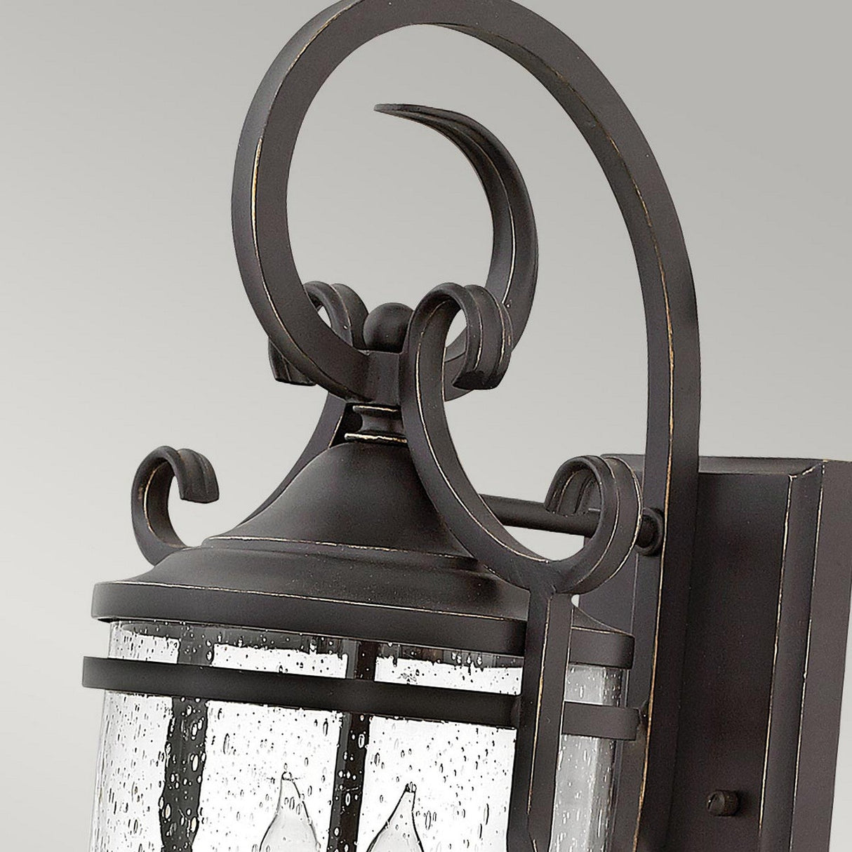 Close-up of the Casa 2 Light Outdoor Wall Lantern - Black, exuding rustic charm with its curved wrought iron design and seeded glass. The dark metal features elegant scrollwork, while the glass has a speckled texture. This charming lantern is mounted on a flat, dark backplate.