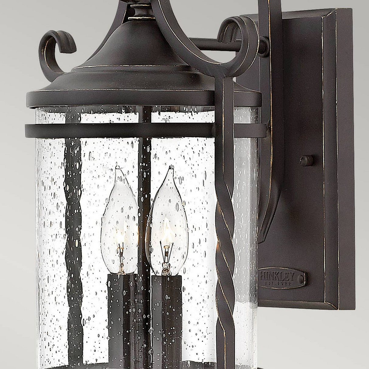 Introducing the Casa 2 Light Outdoor Wall Lantern, which exudes rustic charm with its wrought iron frame and two candle-style bulbs encased within a cylindrical seeded glass enclosure. This fixture boasts a decorative twist design on its arm, finished in sleek black for a striking contrast against its surroundings.