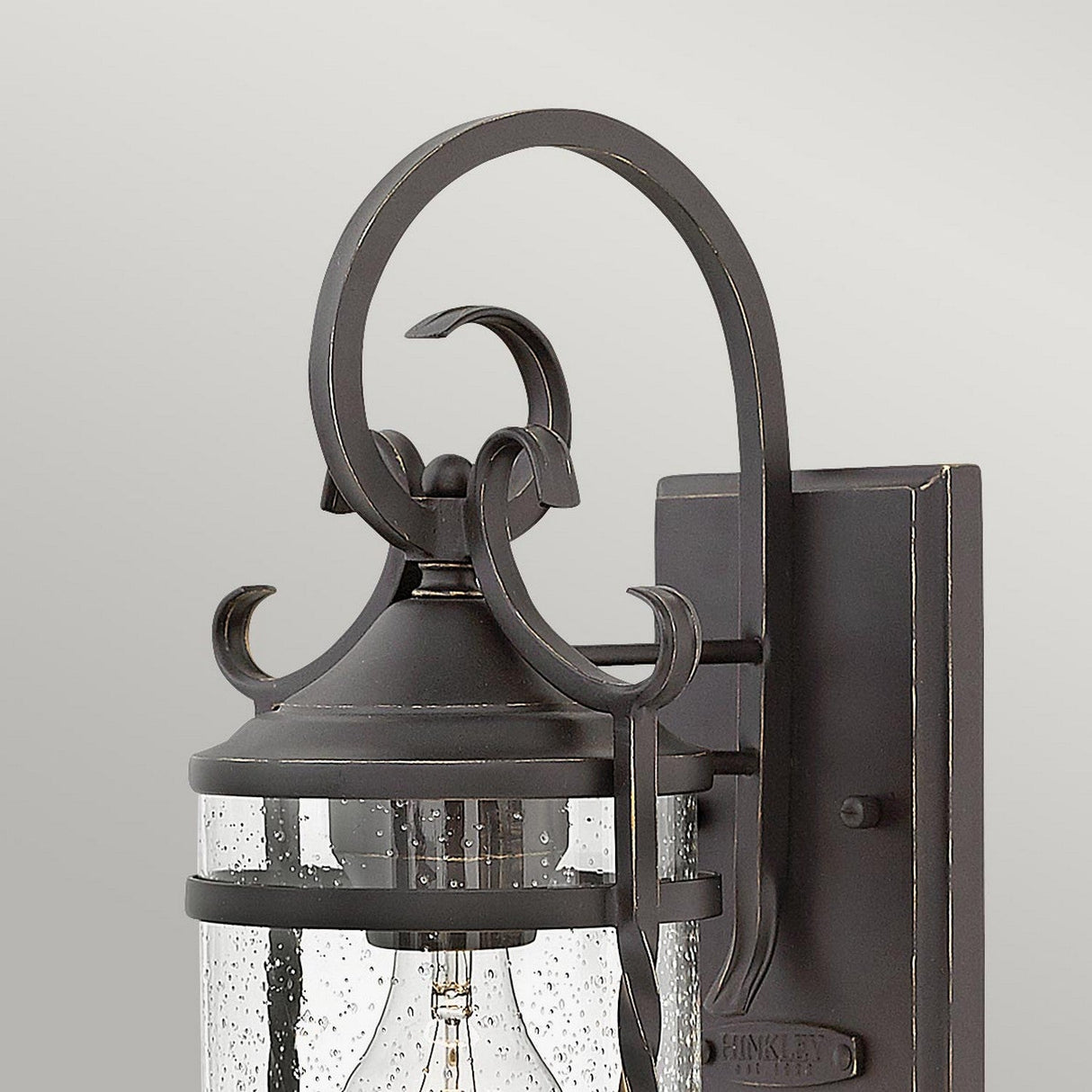 Close-up of the Casa 1 Light Outdoor Wall Lantern in black, showcasing its rustic charm and ornate wrought iron design with a clear glass shade. The lantern is elegantly mounted on a light gray wall, featuring a curved arm and intricate detailing on its rectangular base.