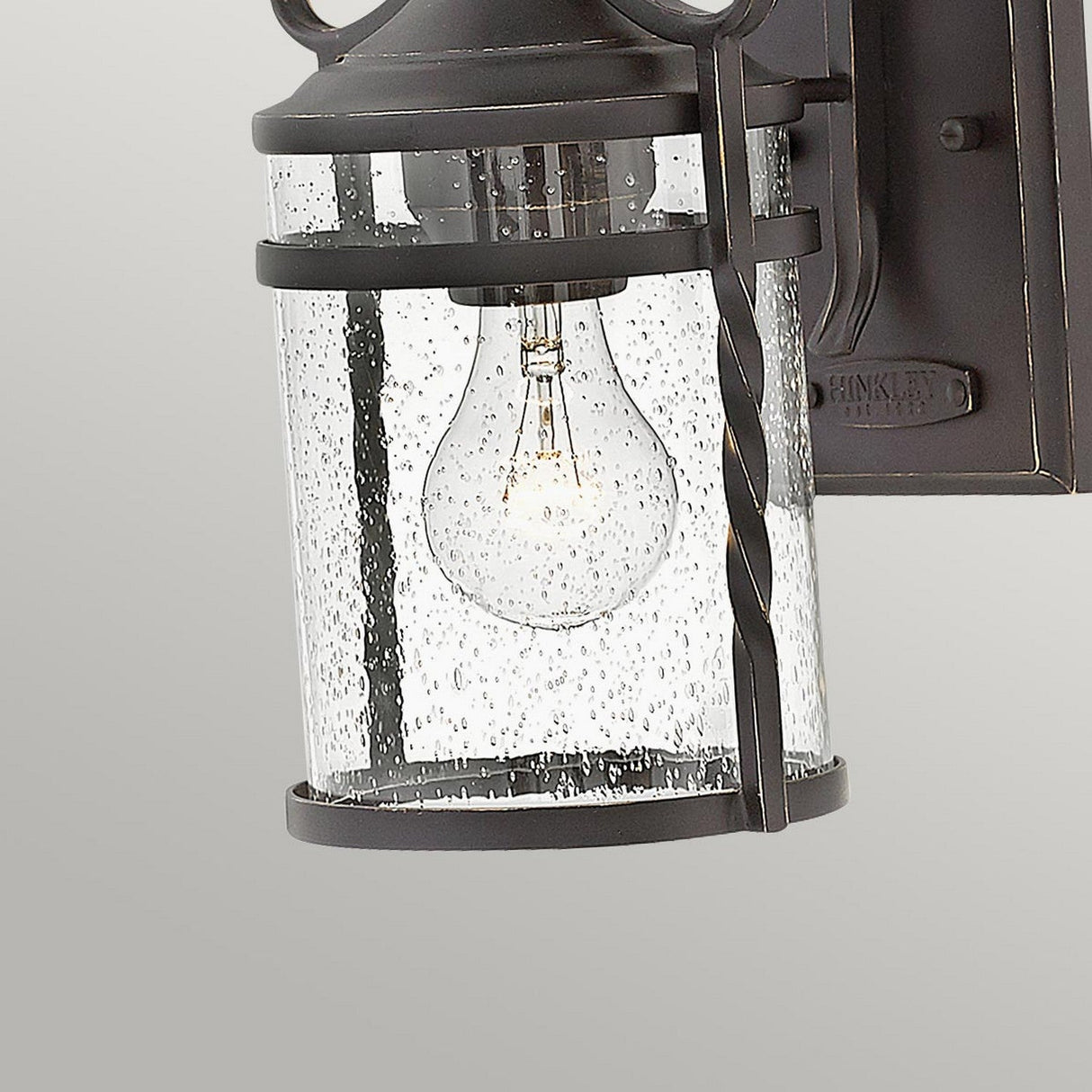 The Casa 1 Light Outdoor Wall Lantern in Black features a cylindrical glass casing embellished with raindrop textures, inside of which a glowing bulb is encased in wrought iron. Set against a light gray background, this lantern exudes rugged charm.