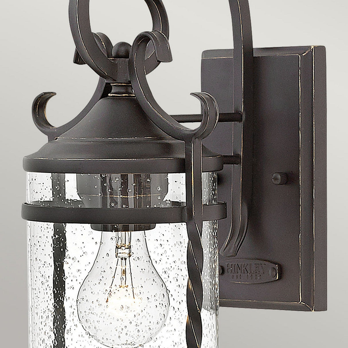 The Casa 1 Light Outdoor Wall Lantern - Black exudes rustic charm with its black wrought iron design. It features a transparent, bubbled glass cover that showcases a visible bulb inside, all elegantly suspended from a decorative curved metal bracket.