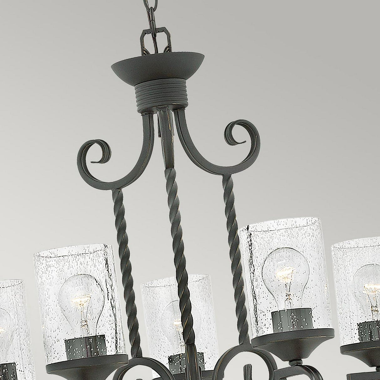 A close-up view of the Casa 5 Light Chandelier - Black highlights its elegant twisted metal arms and distinctive seedy glass shades. The wrought iron-framed shades feature a speckled texture that beautifully reveals the exposed bulbs within, all set against a subtle light gray background.