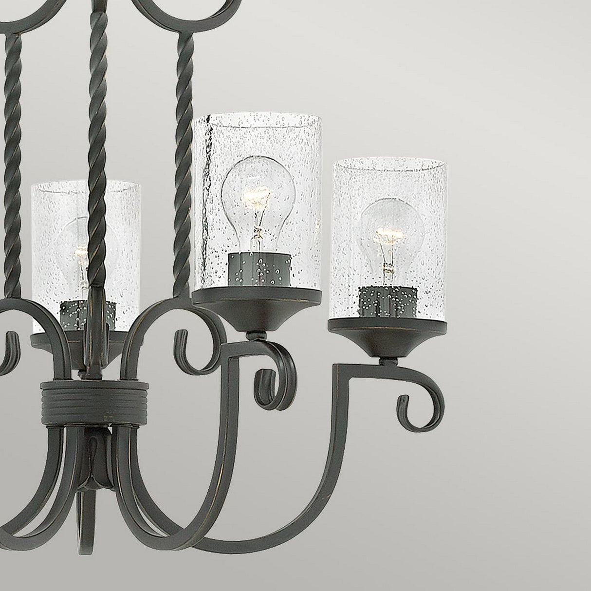 A close-up view of the Casa 5 Light Chandelier - Black showcases its elegant wrought iron frame adorned with intricate scroll designs. It features three seedy glass shades, each housing a light bulb, set against a plain, light gray background.