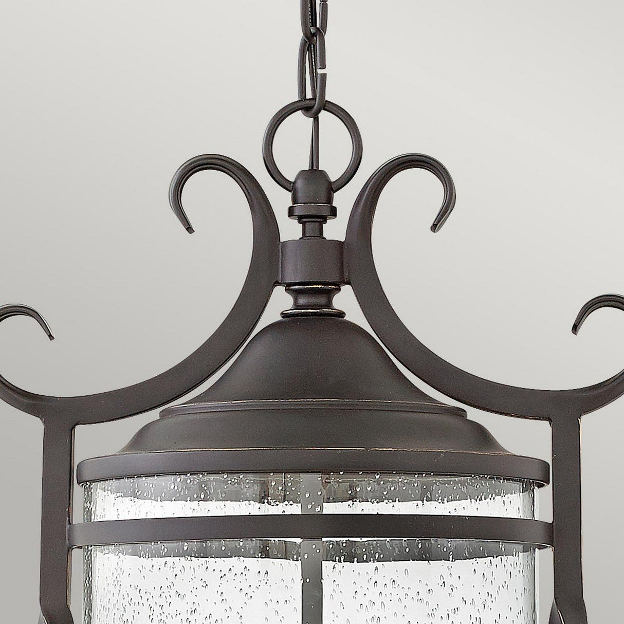 A close-up of the Casa 3 Light Outdoor Chain Lantern - Black, showcasing its ornate design. The lantern features clear seedy glass panels and elegant scrollwork details, highlighted by intricate pierced metalwork on top, hanging against a light gray background.