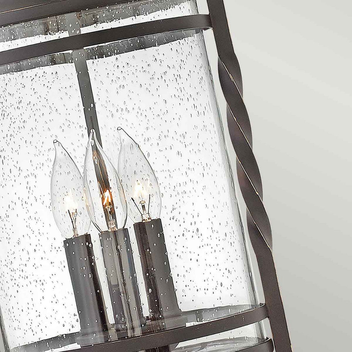 The Casa 3 Light Outdoor Chain Lantern - Black showcases a close-up view of its design, featuring three candle-shaped bulbs within a clear seedy glass cylinder. It is encased in a pierced metalwork frame boasting a dark finish and a twisted design element on the side.