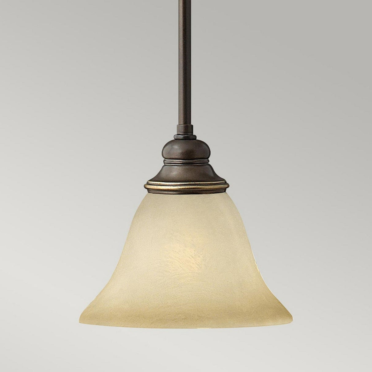 The Cello 1 Light Mini Pendant in antique bronze showcases a frosted glass bell-shaped shade that exudes the charm of vintage faux alabaster glass set against a plain gray background.