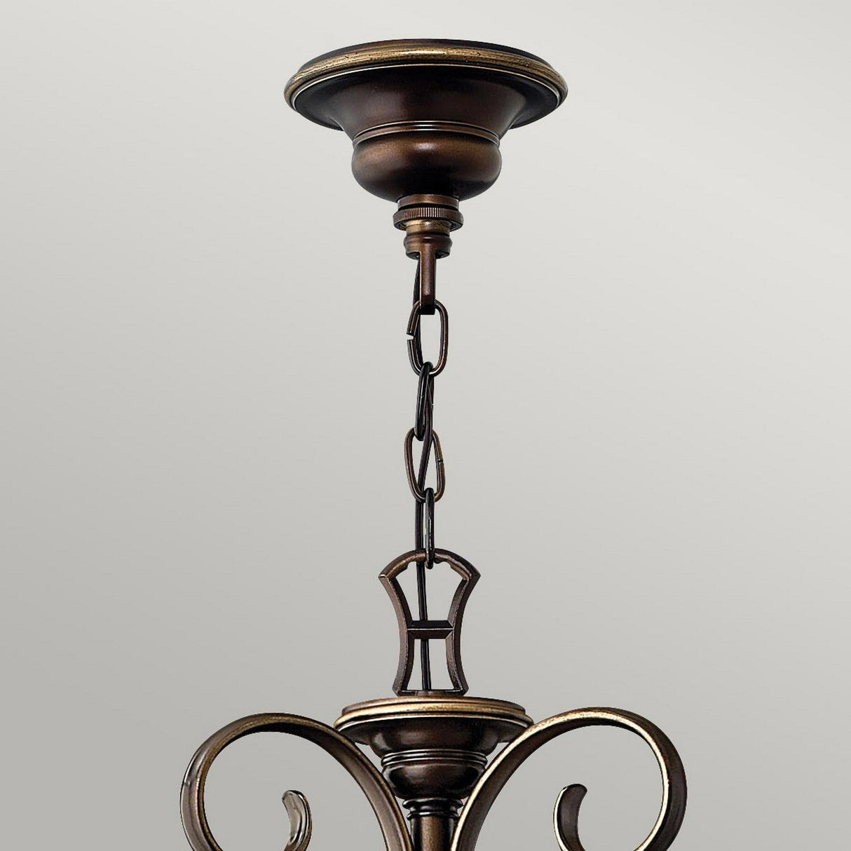 The Cello 15 Light Chandelier in Antique Bronze boasts an intricate, curved design complemented by vintage-style faux alabaster glass. It is suspended elegantly from a chain linked to a decorative ceiling mount. The image emphasizes the upper portion of the chandelier against a plain backdrop.