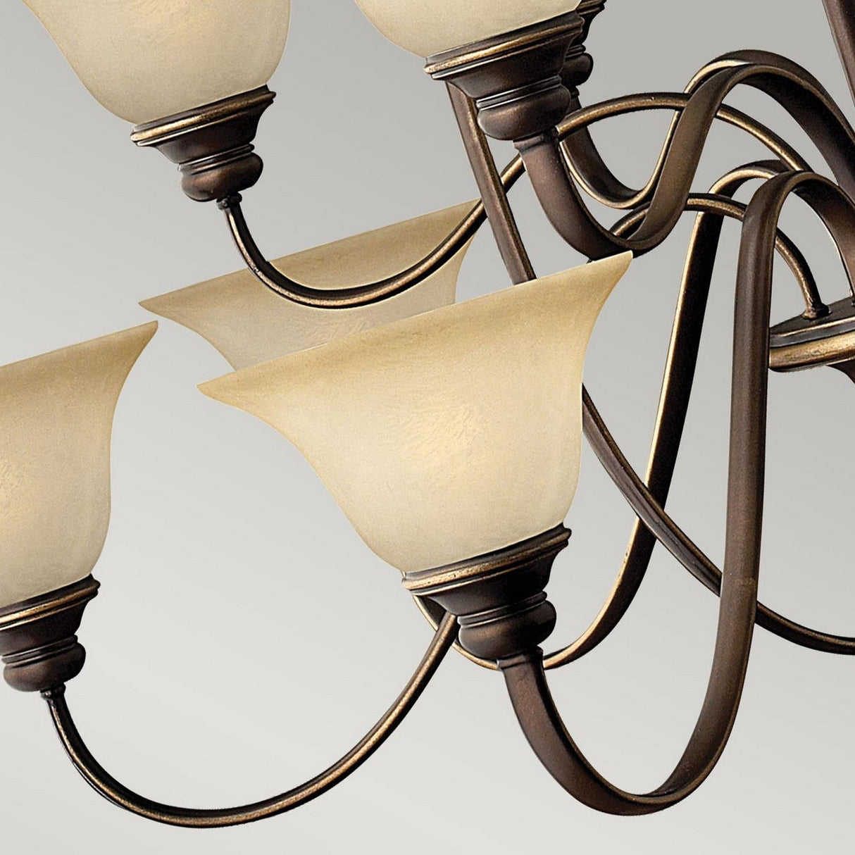 Close-up of the Cello 15 Light Chandelier in Antique Bronze, featuring ornate curved arms and vintage faux alabaster glass shades, set against a neutral gray background. The elegant design highlights swirling metalwork and warm-toned light fixtures.