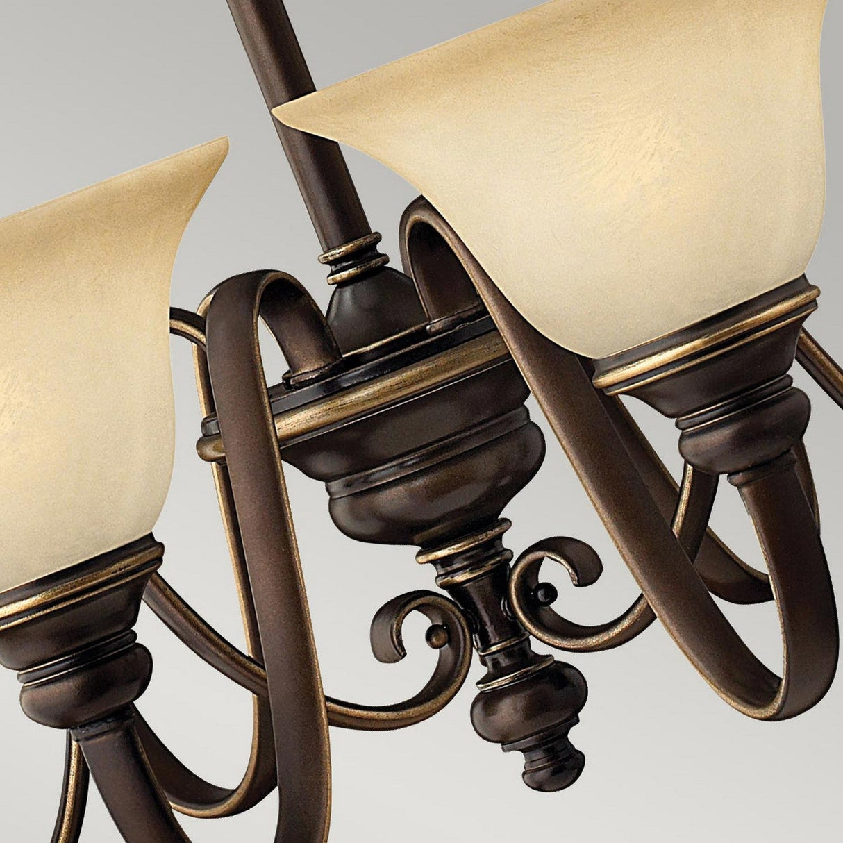Close-up of the Cello 6 Light Chandelier in antique bronze, featuring intricate swirling metal arms and two upward-facing shades made from vintage alabaster cream-colored glass, set against a gray background.