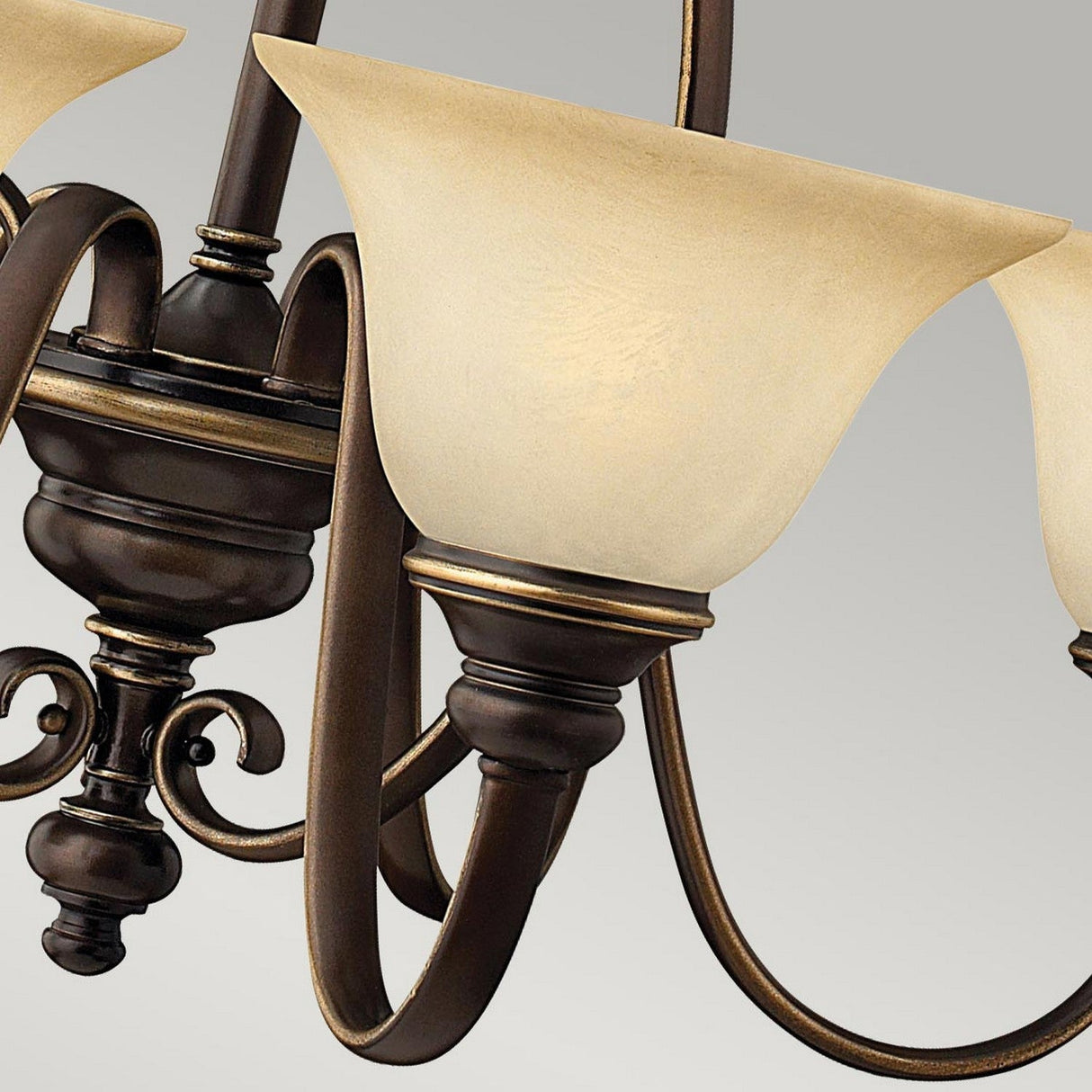The Cello 6 Light Chandelier in Antique Bronze showcases a detailed close-up of its bronze-finished metal arms and intricate scrollwork. Adorned with vintage alabaster glass lampshades, it casts a warm, soft glow against a plain gray background.