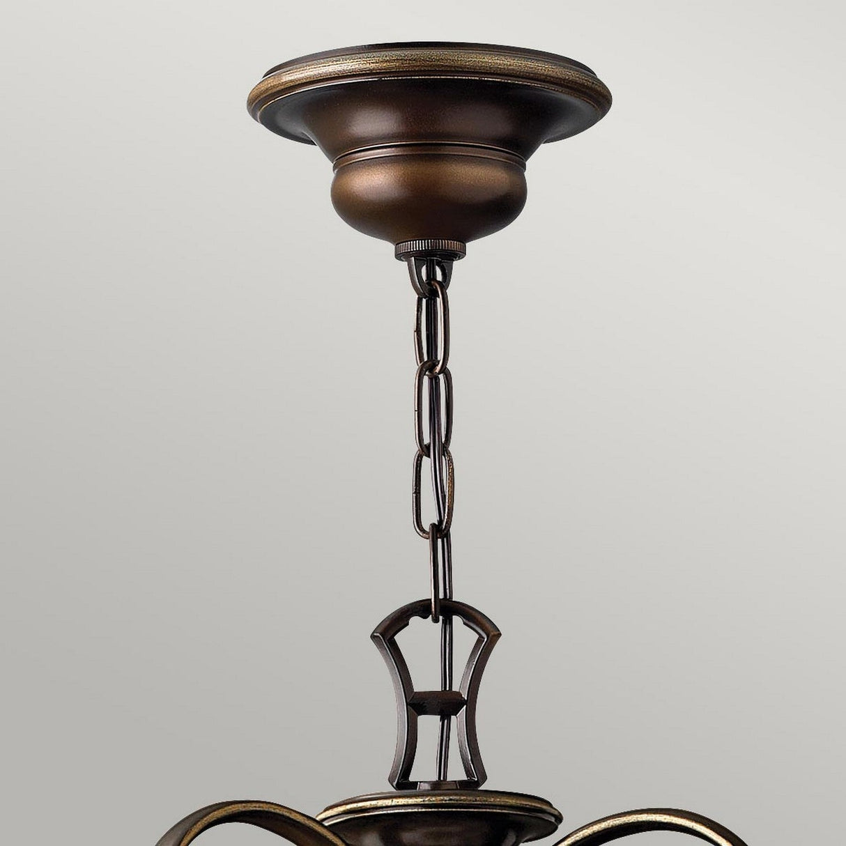 A close-up of the Cello 9 Light Chandelier in antique bronze features its bronze chain and mounting fixture, highlighting an elegant metal canopy with antique bronze detailing against a light gray background, complemented by the soft glow of faux alabaster glass.