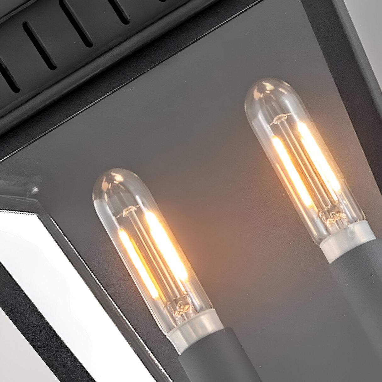 Close-up of two lit LED filament bulbs inside the Chapel Hill 2 Light Outdoor Half Wall Lantern in black. The glass-enclosed fixture emits a warm, yellowish glow for stylish outdoor lighting.