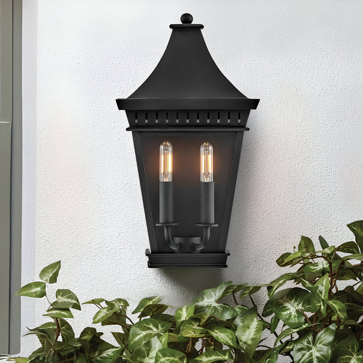 The Chapel Hill 2 Light Outdoor Half Wall Lantern in black is mounted on a white textured wall above green leafy plants.